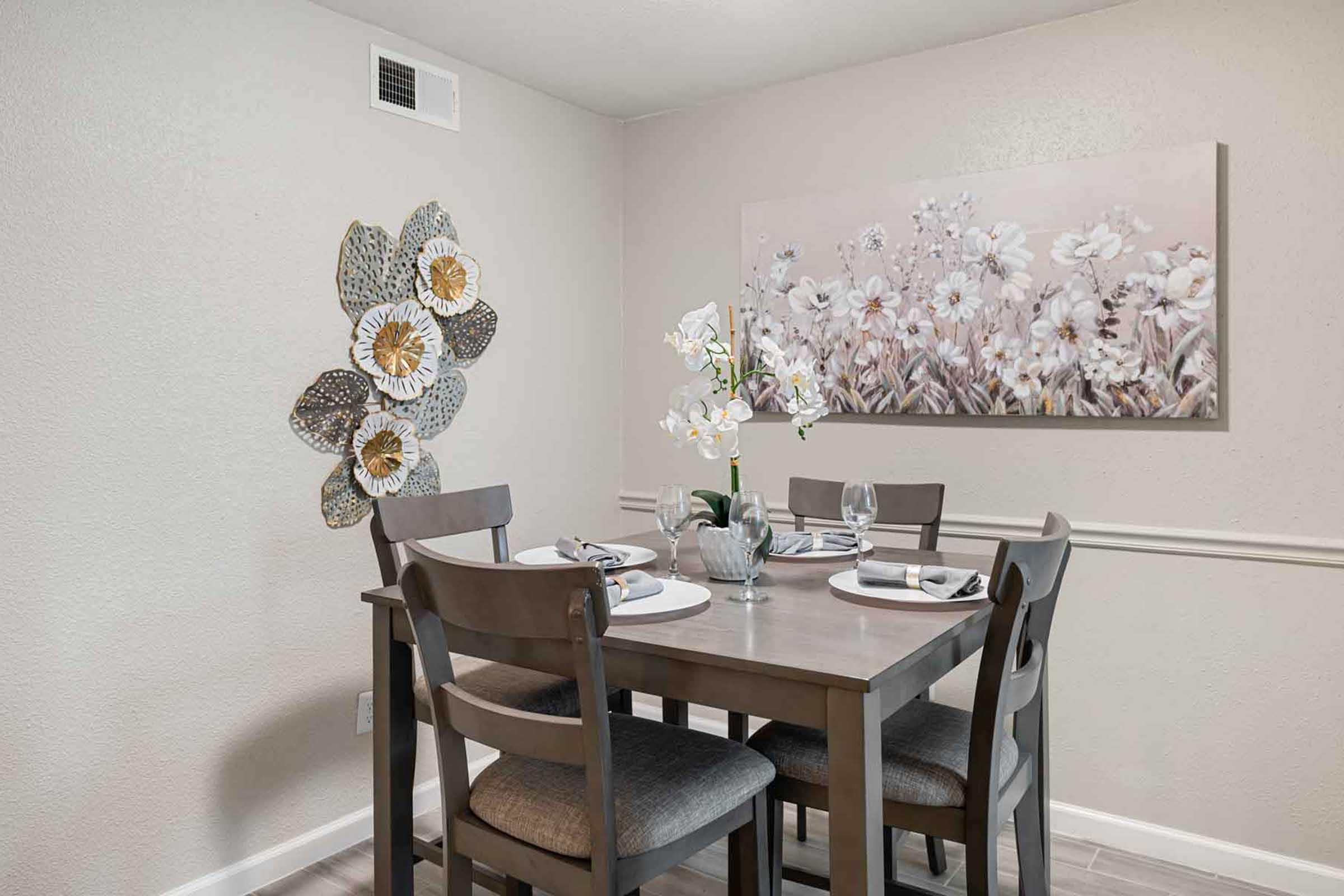 A cozy dining area featuring a rectangular gray table set for four with plates, glasses, and napkins. The walls are painted a light color, adorned with a floral artwork and decorative wall accents resembling flowers. Natural light enhances the welcoming ambiance.