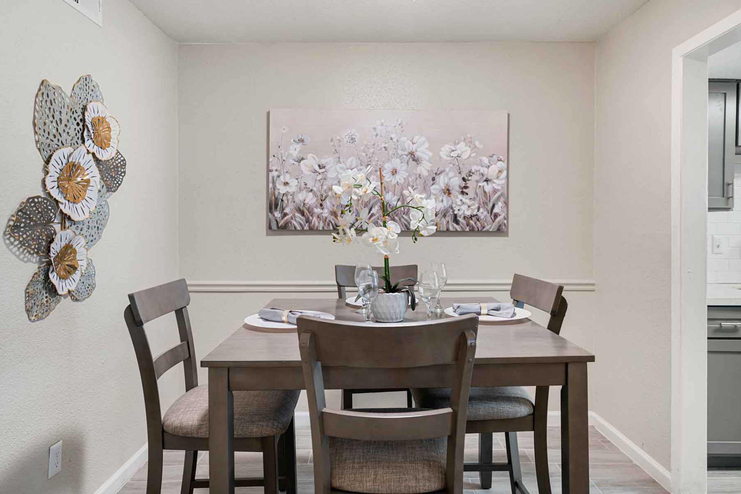 A modern dining area featuring a round wooden table set for four, with elegant place settings. A floral painting hangs on the wall, complemented by a decorative wall piece made of leaves and flowers. The room has soft, neutral colors, creating a warm and inviting atmosphere.