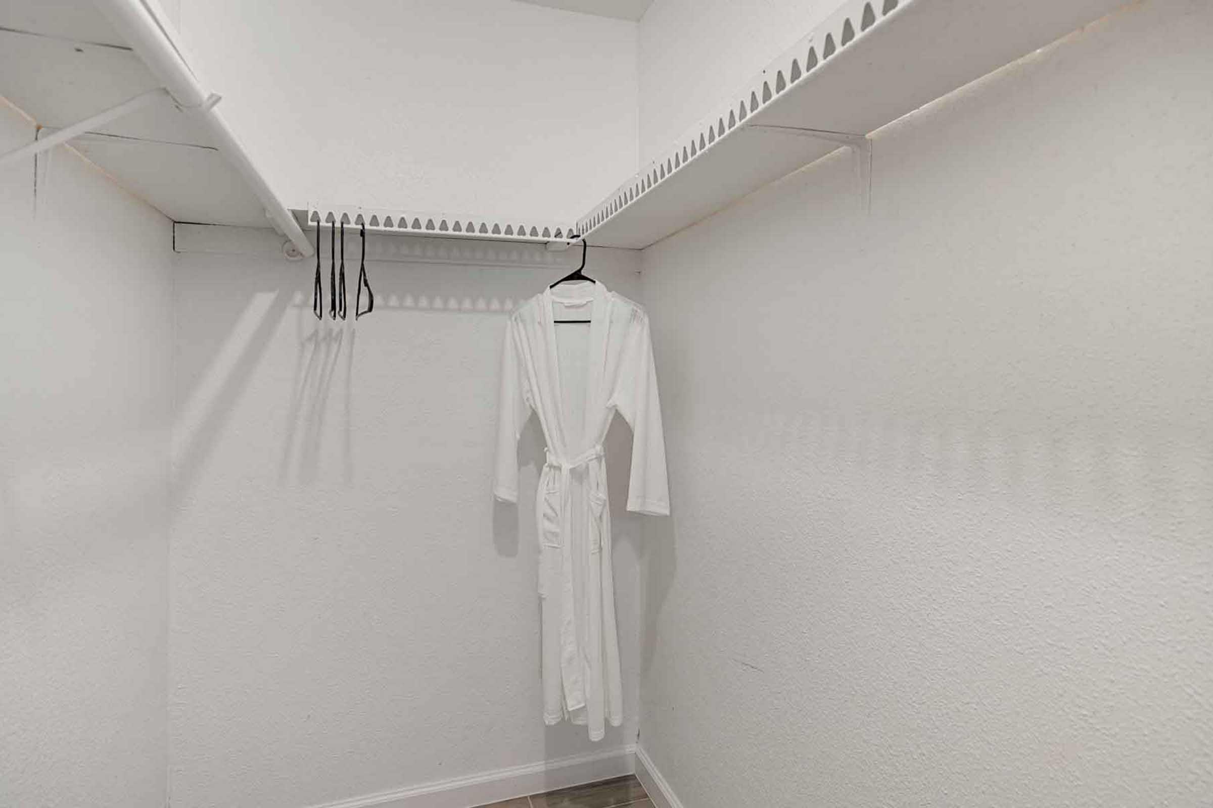 A minimalistic closet featuring white walls and a wooden floor. A white robe hangs on a hanger from one of the shelves. There are additional hangers and a simple, organized layout with ample space.