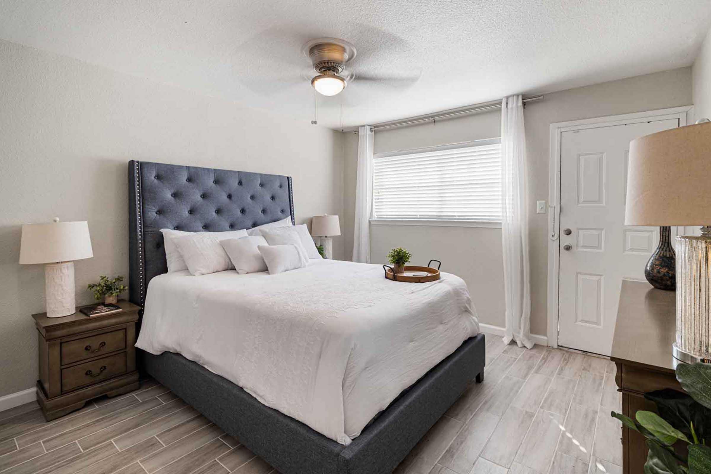 Bright and airy bedroom featuring a cozy, tufted headboard, white bedding, and decorative pillows. Natural light streams through a window dressed with sheer curtains. A bedside table holds a small plant and a tray. The room includes a ceiling fan, wooden flooring, and a door leading outside.