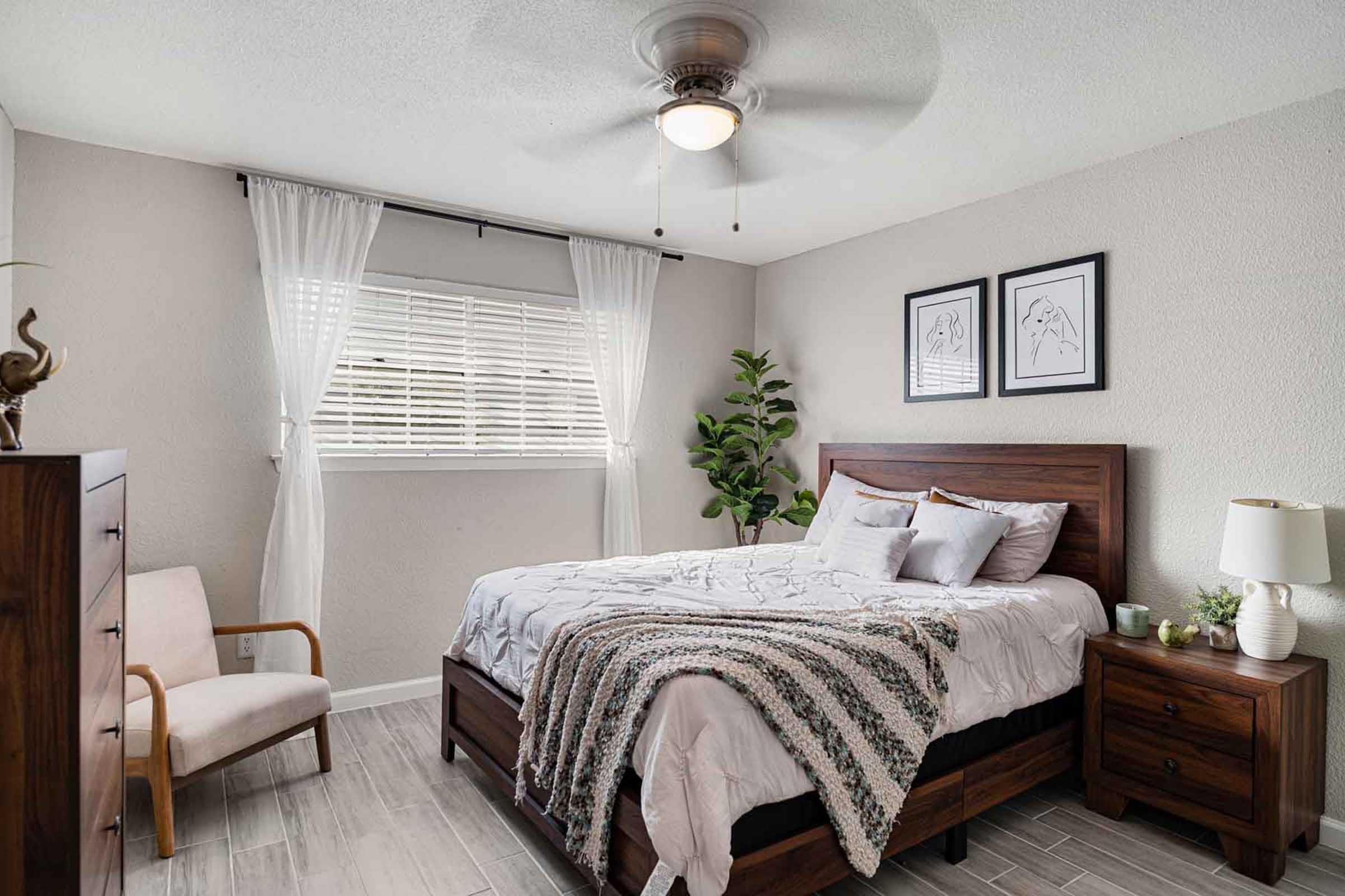 A cozy bedroom featuring a wooden bed with soft bedding, a bedside table with a lamp, and a modern chair. Natural light filters through the window adorned with sheer curtains. The walls are decorated with framed artwork, and a decorative plant adds a touch of greenery to the space.