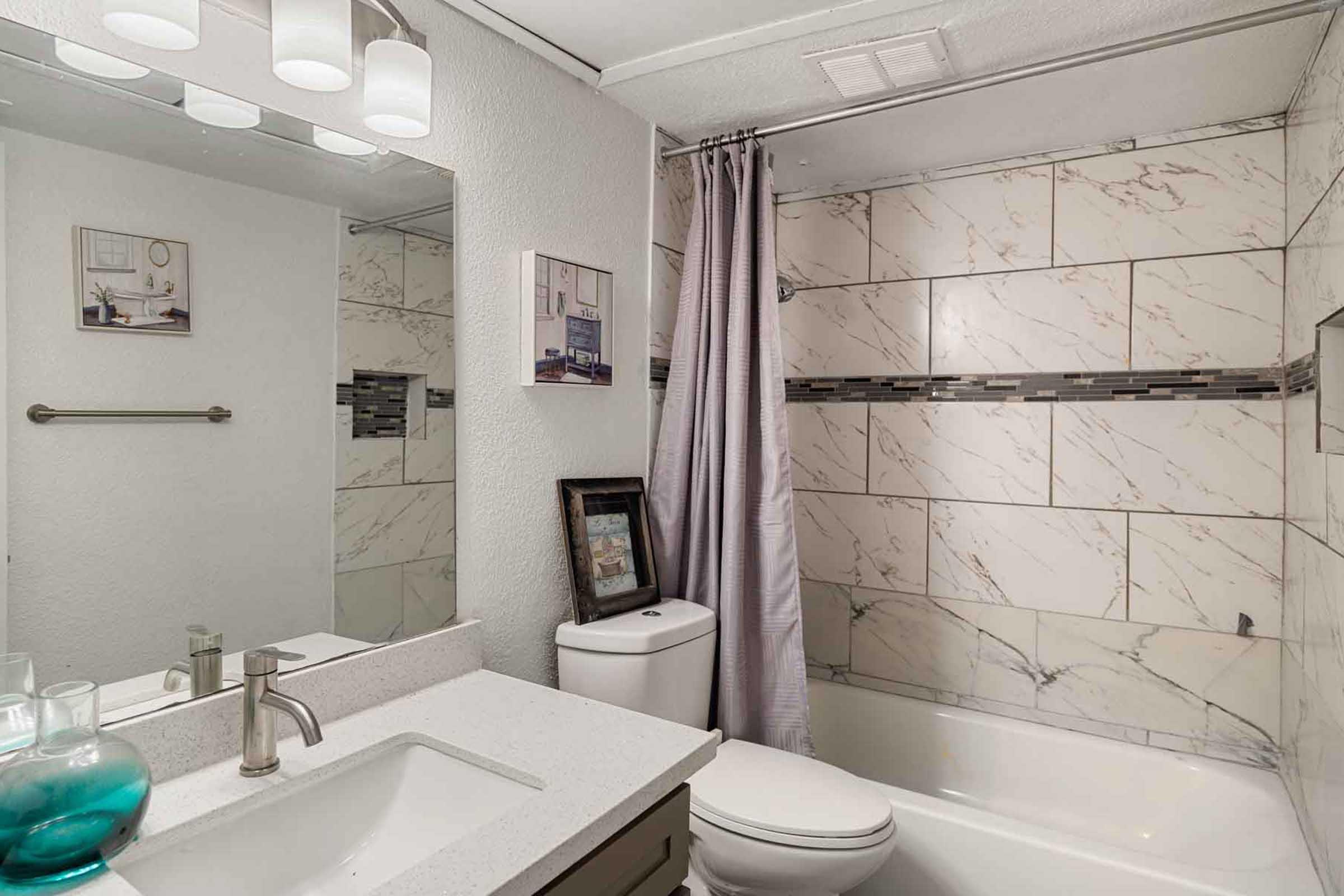 Modern bathroom featuring a bathtub with a shower curtain, marble-patterned tiles, a sleek vanity with a sink, and a mirror above it. Light fixtures illuminate the space, and a decorative picture frame is placed on a shelf, adding a touch of decor.