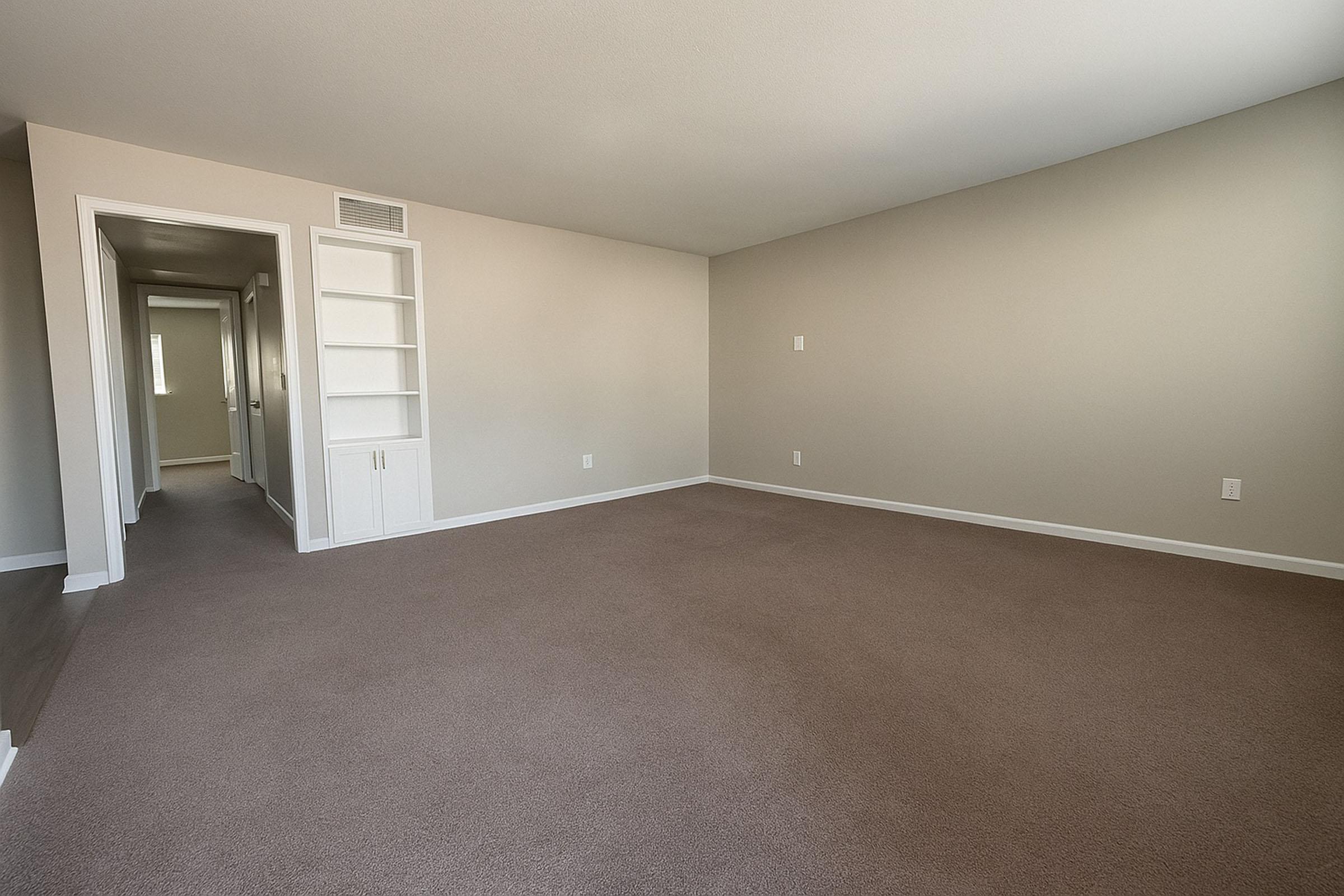 A spacious, empty room with light beige walls and carpet. There is a white built-in shelving unit on the left, and a hallway leading to another room is visible in the background. Natural light may come through windows, enhancing the room’s neutral tone. Ideal for various uses or setups.