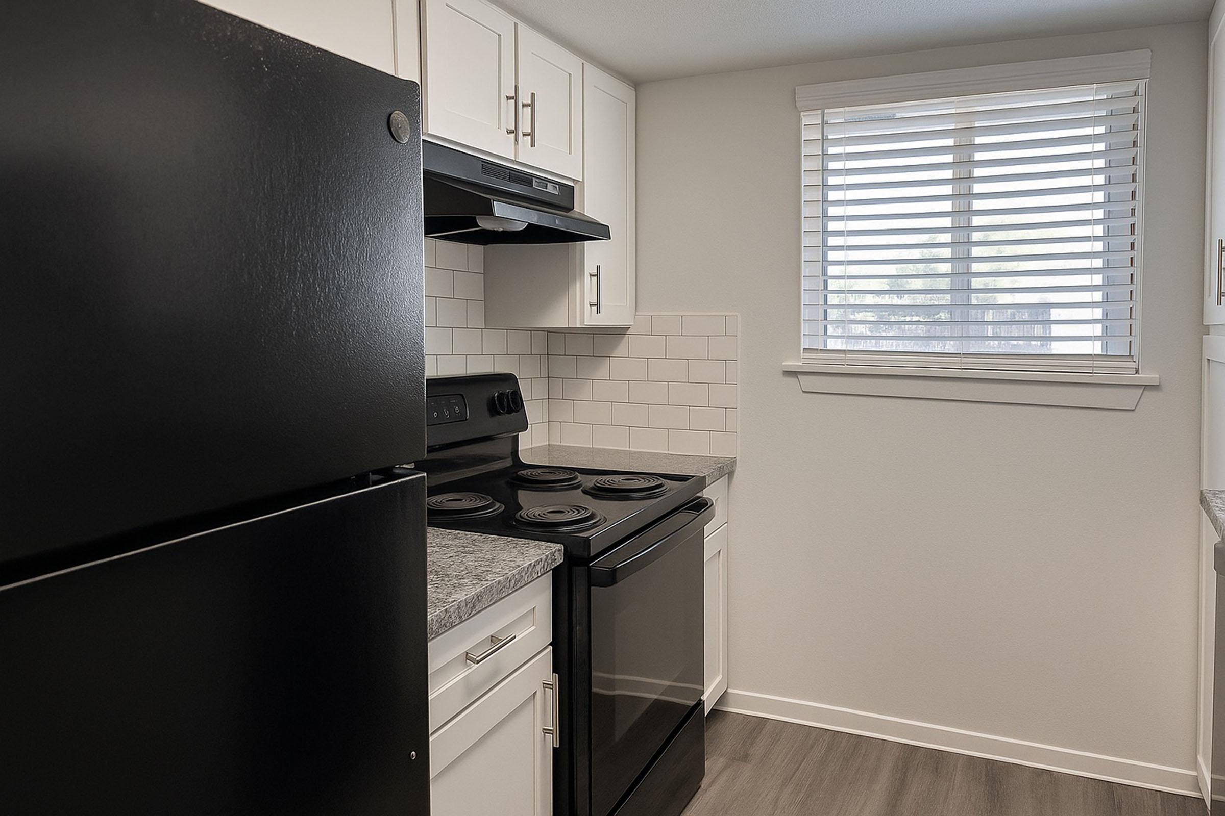 A modern kitchen featuring a black refrigerator and an electric stove with four burners. The cabinetry is white, complemented by a light gray countertop. There is a window with white blinds, allowing natural light to illuminate the space, and light-colored flooring adds to the bright atmosphere.