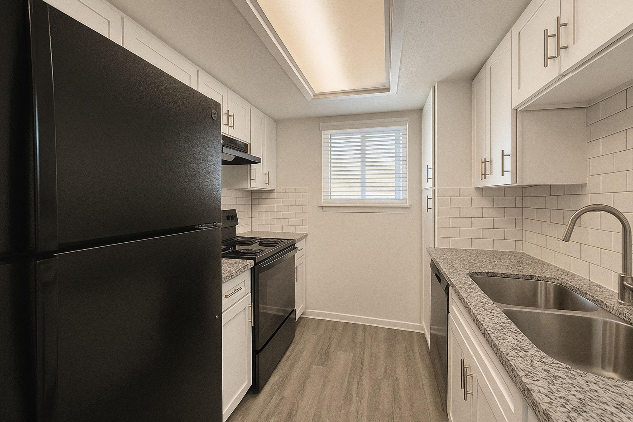 A modern kitchen featuring black appliances, including a refrigerator and stove, sleek white cabinetry, and gray countertops. The space has a window with blinds, allowing natural light, and light-colored flooring. The design is clean and functional, ideal for contemporary living.