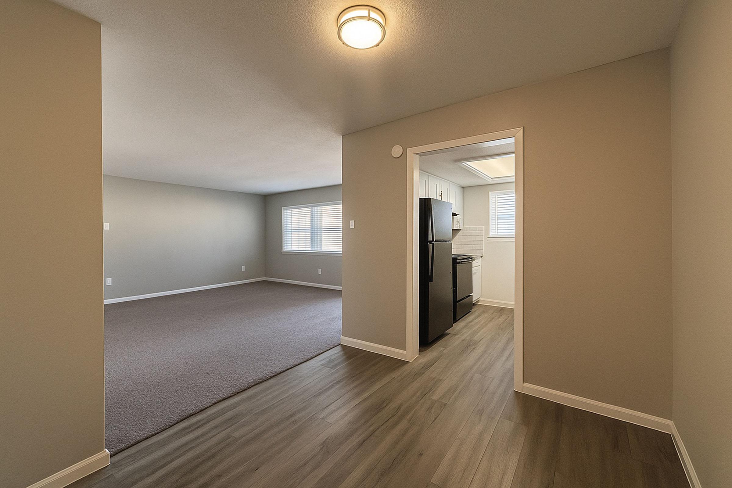 A spacious interior view of an apartment featuring a light-colored wall and flooring. The area includes a carpeted living room leading to a kitchen with modern appliances visible through an open doorway. The room is well-lit with natural light from windows.