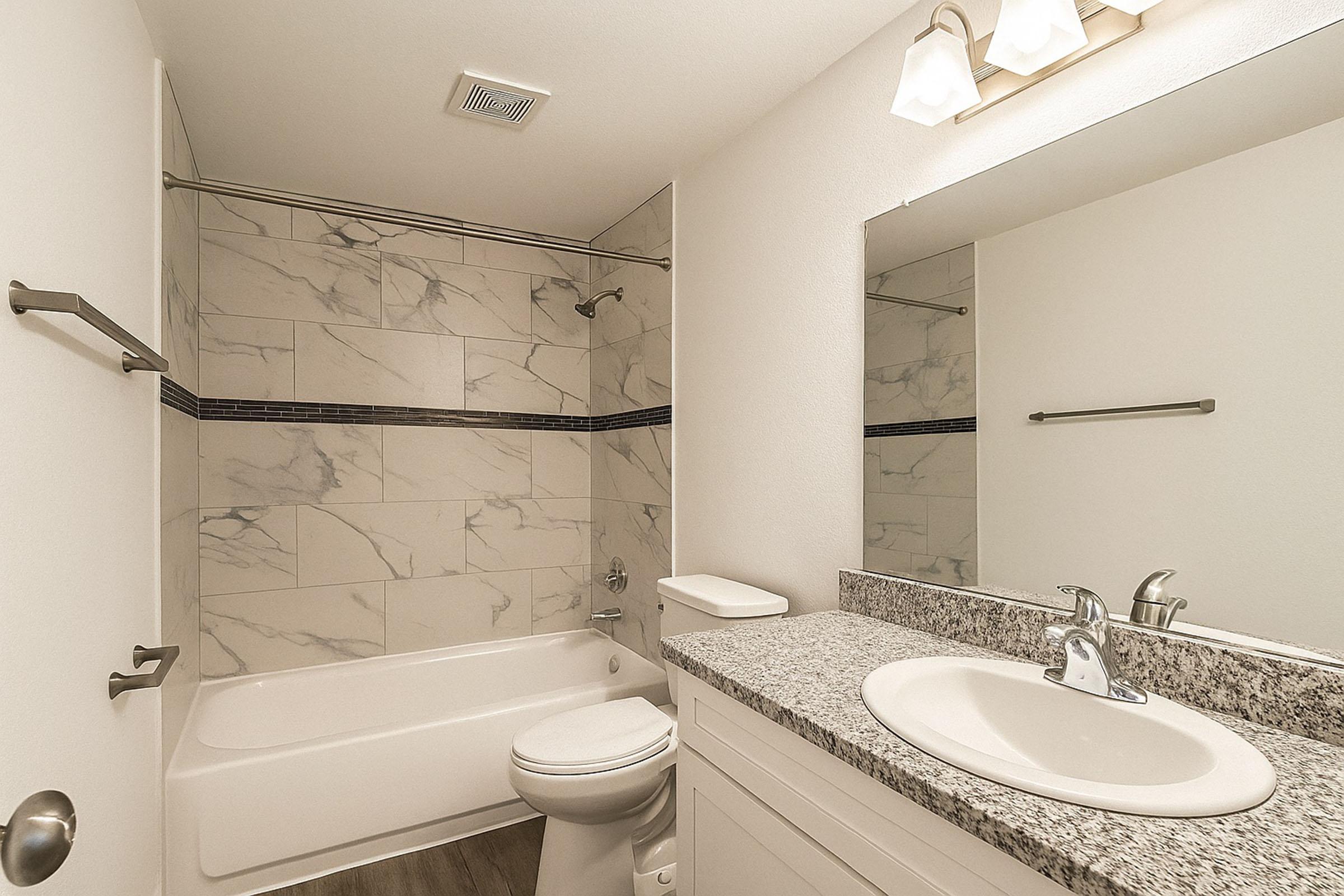 A modern bathroom featuring a bathtub with a shower, marble tiled walls, a granite countertop with a sink, and a large mirror above. The space includes a toilet and a light fixture with three bulbs. The overall design is clean and contemporary with neutral colors.