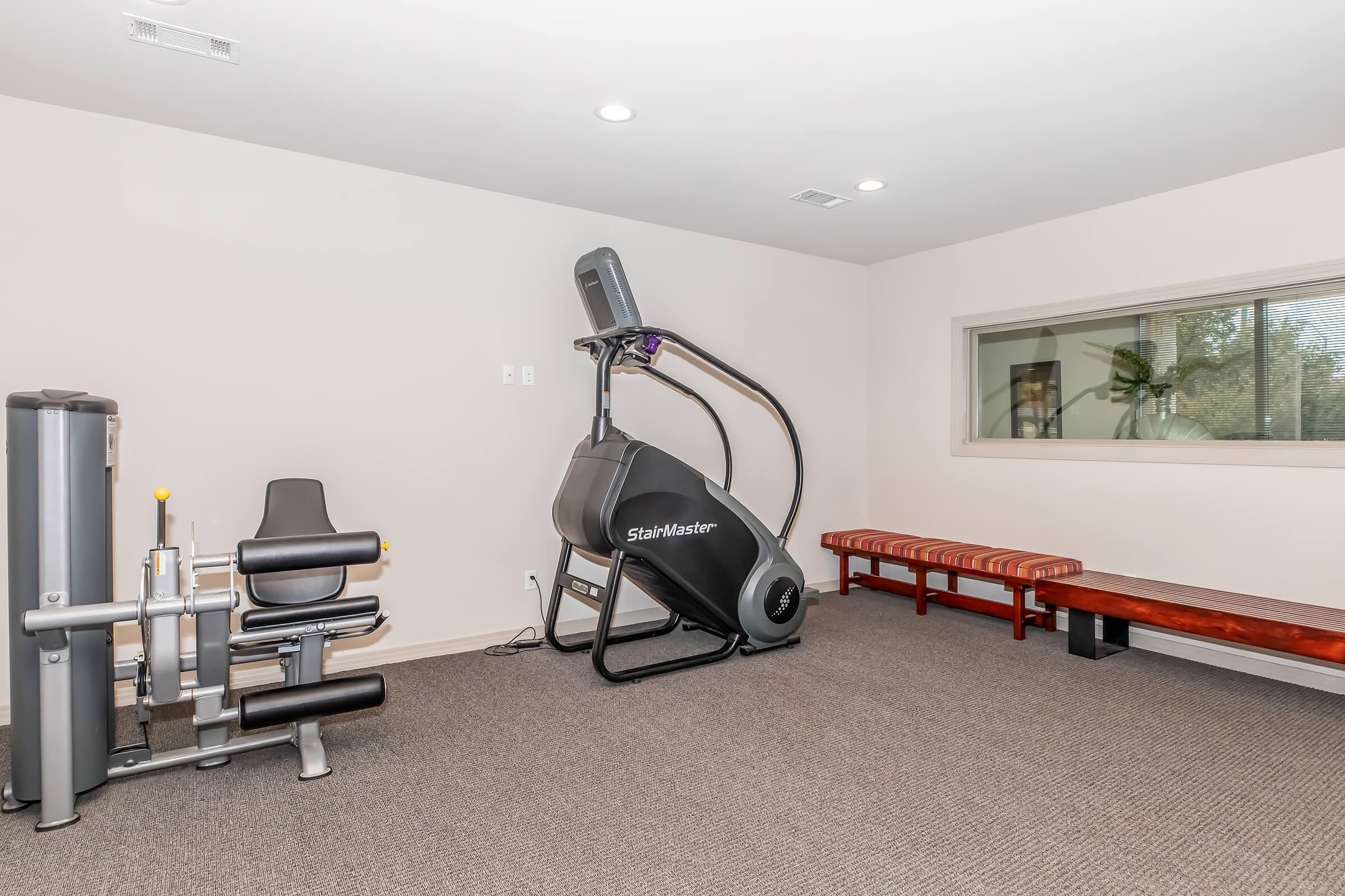 A modern gym interior featuring a StairMaster exercise machine, a weight machine, and a bench, all set against a neutral-colored wall. Large window allows natural light into the space, highlighting the clean and spacious layout designed for fitness activities.