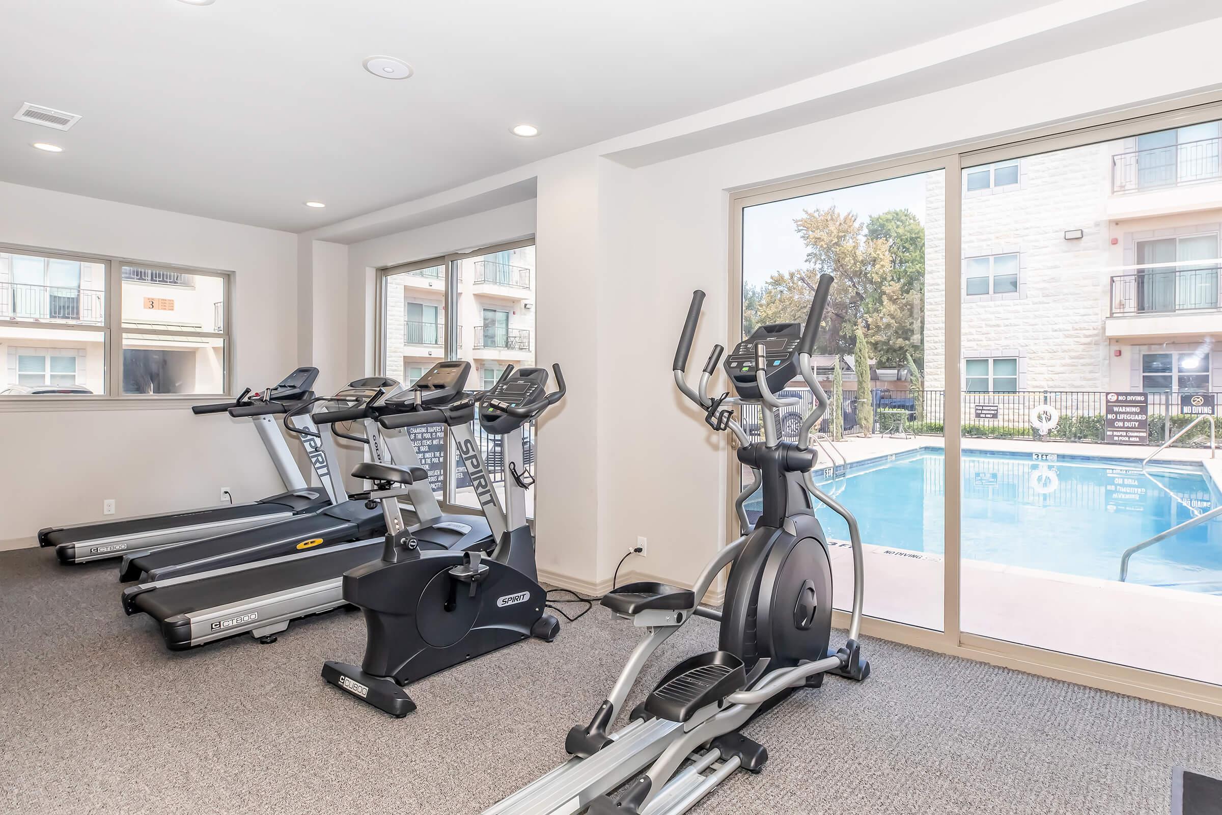 Fitness center with several exercise machines, including treadmills and an elliptical, overlooking a pool area through large windows. The space has carpet flooring and natural light streaming in.
