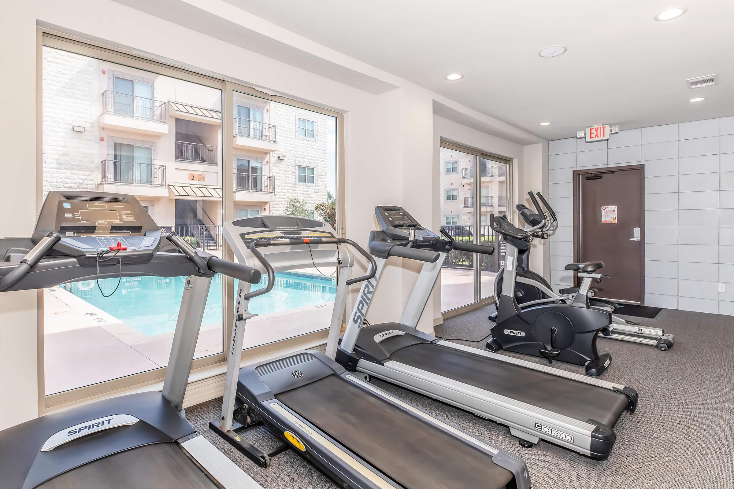 A modern fitness room featuring several machines, including treadmills and an elliptical trainer, with large windows overlooking a swimming pool. The space is well-lit and has a clean, contemporary design.