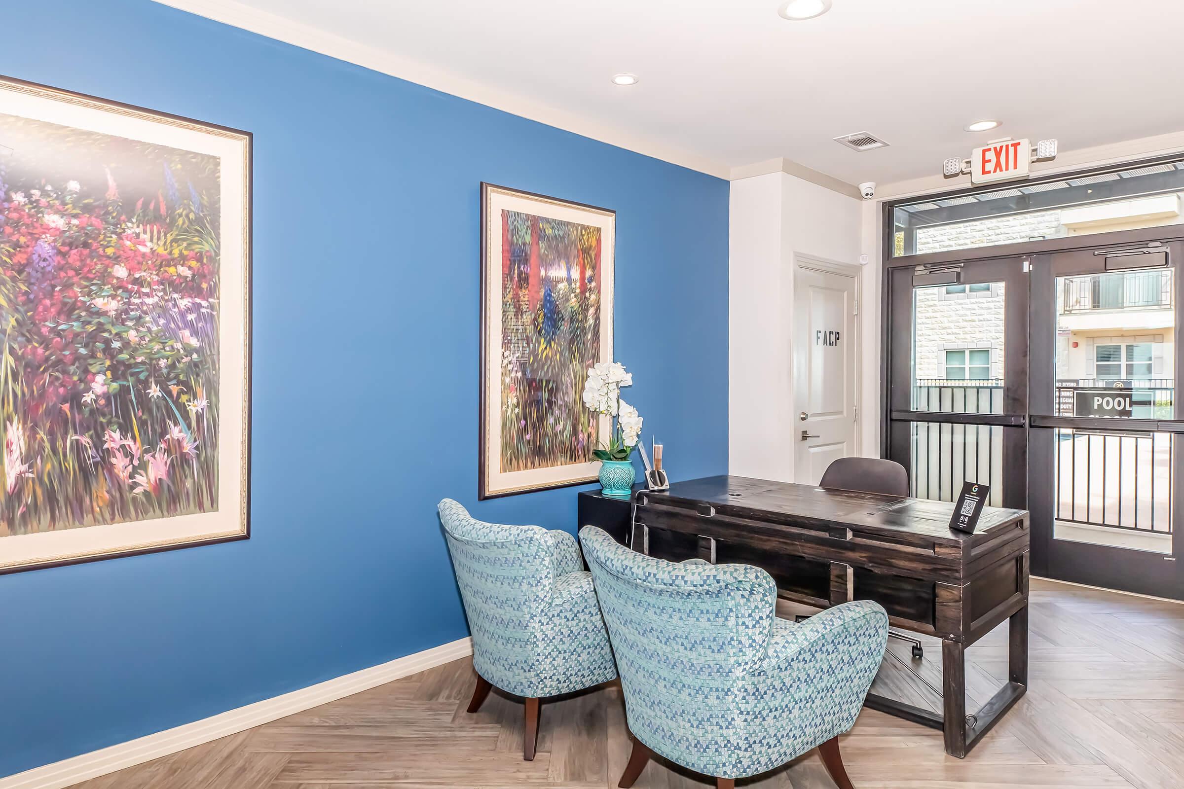 A modern reception area featuring a blue accent wall, two patterned armchairs, a wooden desk with a phone, and a decorative vase with an orchid. Colorful floral artwork hangs on the walls, creating a welcoming atmosphere. Large glass doors lead to an outdoor pool area.