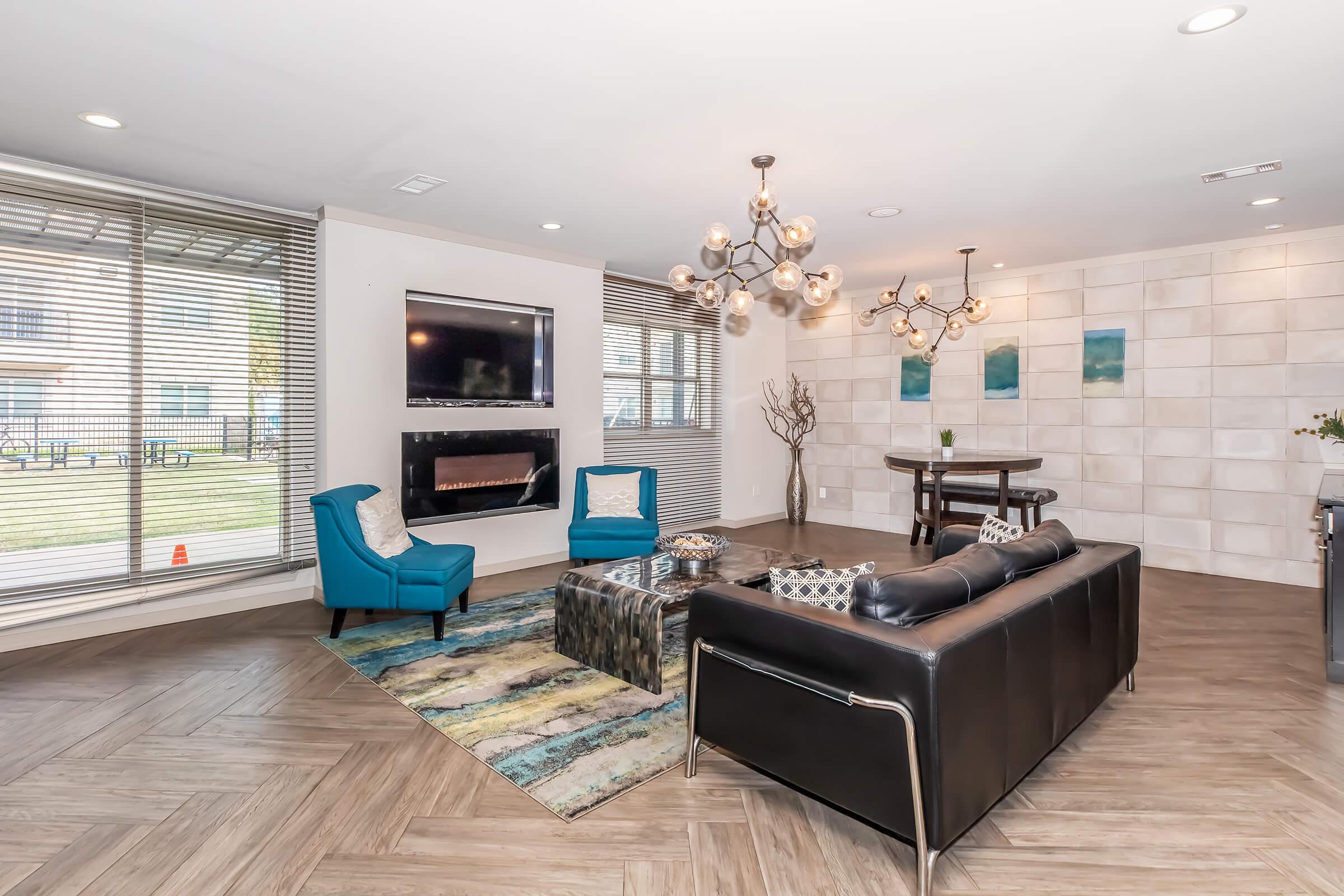 A modern living room featuring two blue chairs, a black leather sofa, and a stylish coffee table on a textured rug. The room has large windows with blinds, a flat-screen TV mounted on the wall, and a contemporary chandelier, creating a bright and inviting atmosphere.