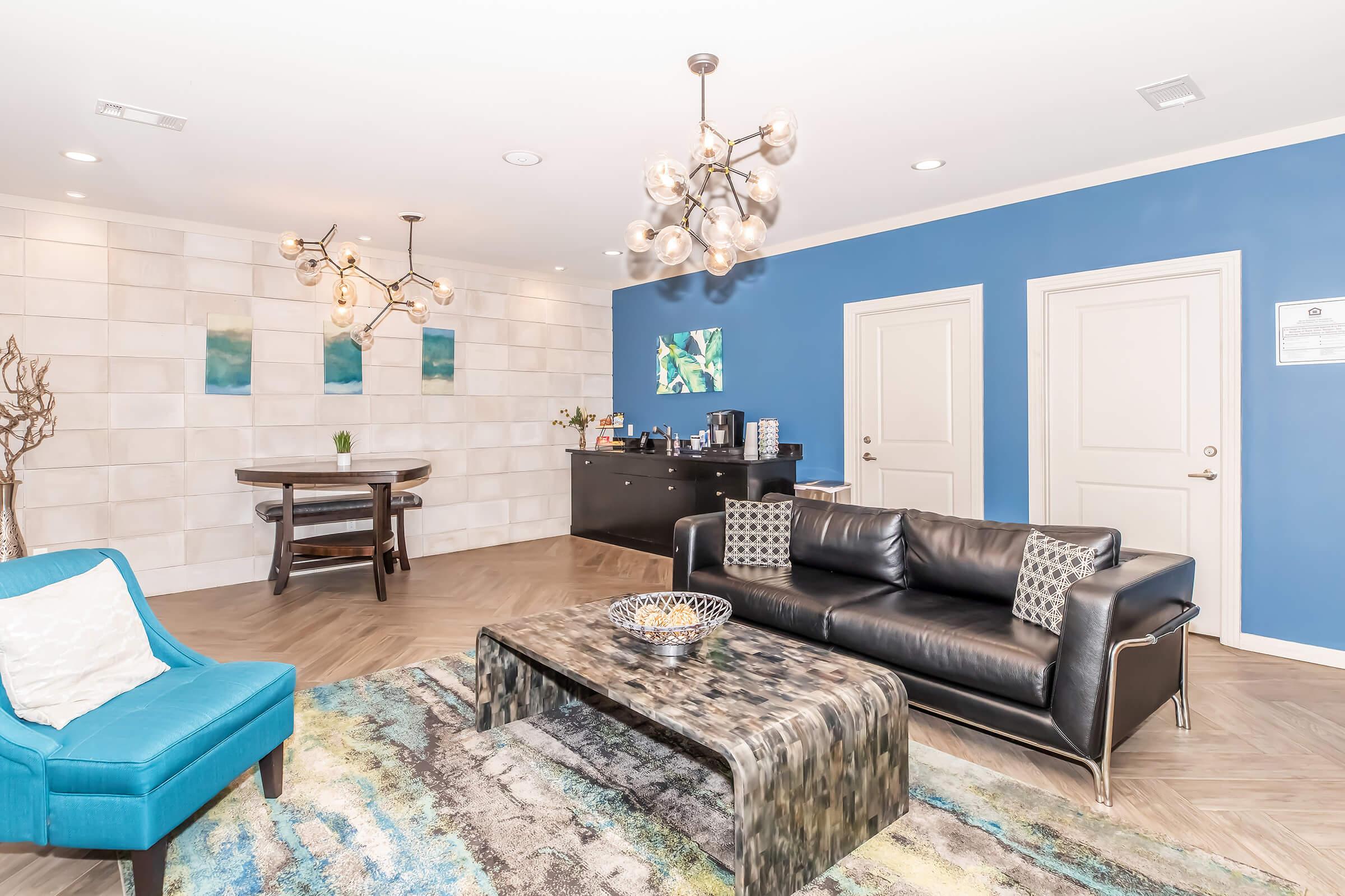 A modern lounge area featuring a black leather sofa with decorative pillows, a stylish coffee table, and a blue accent chair. The walls are a mix of light and dark blue, with abstract art and a round pendant light hanging from the ceiling. There’s a small dining table and a coffee station against the back wall.
