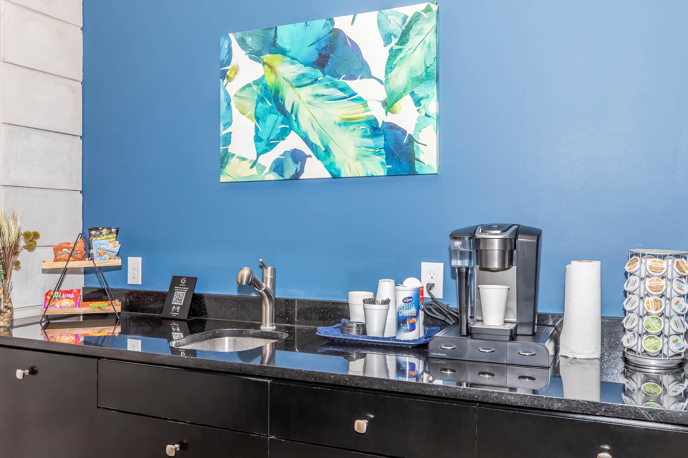 A modern coffee station featuring a coffee maker, cups, and a variety of coffee pods. The station is set against a blue wall with a tropical leaf painting above. A small shelf holds snacks, and there are paper towels nearby. The countertop is sleek and dark, adding to the contemporary feel of the space.
