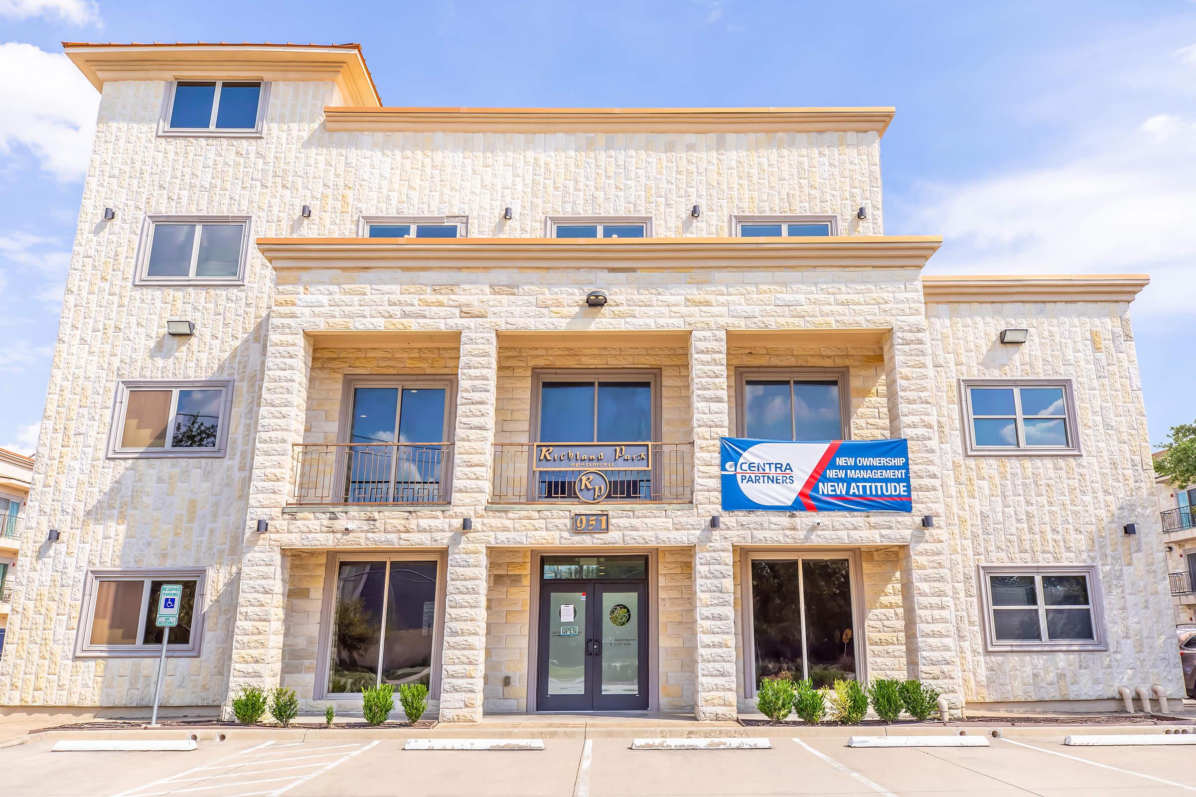 A three-story building made of light-colored stone, featuring large windows and a balcony on the second floor. The entrance has double doors, and there are banners hanging on the front, promoting "Central Painters" and a "New Attitude." There are small shrubs flanking the entrance and parking spaces in front.