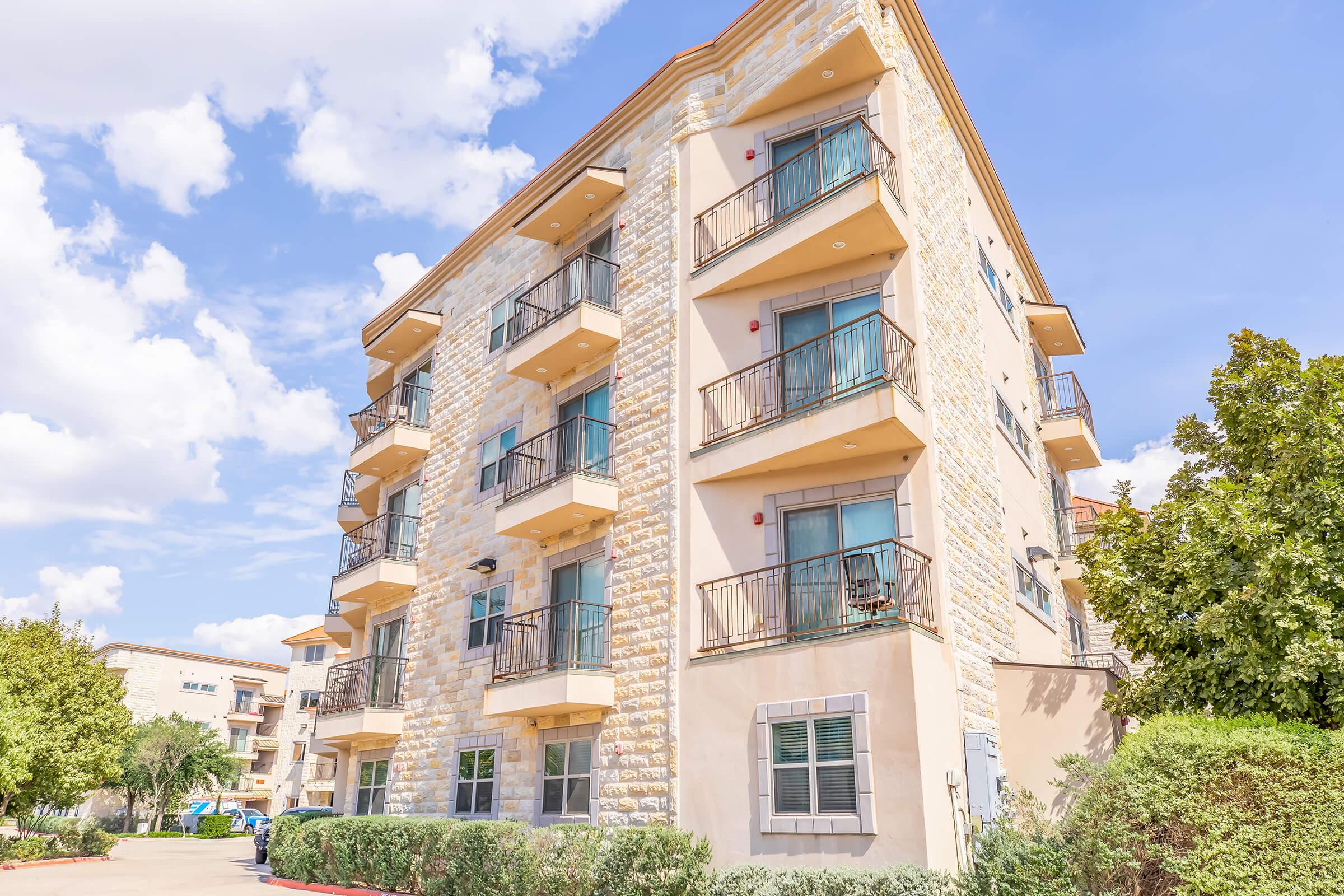 A modern, multi-story apartment building with beige stone exterior, featuring several balconies, green landscaping, and a clear blue sky with scattered clouds.