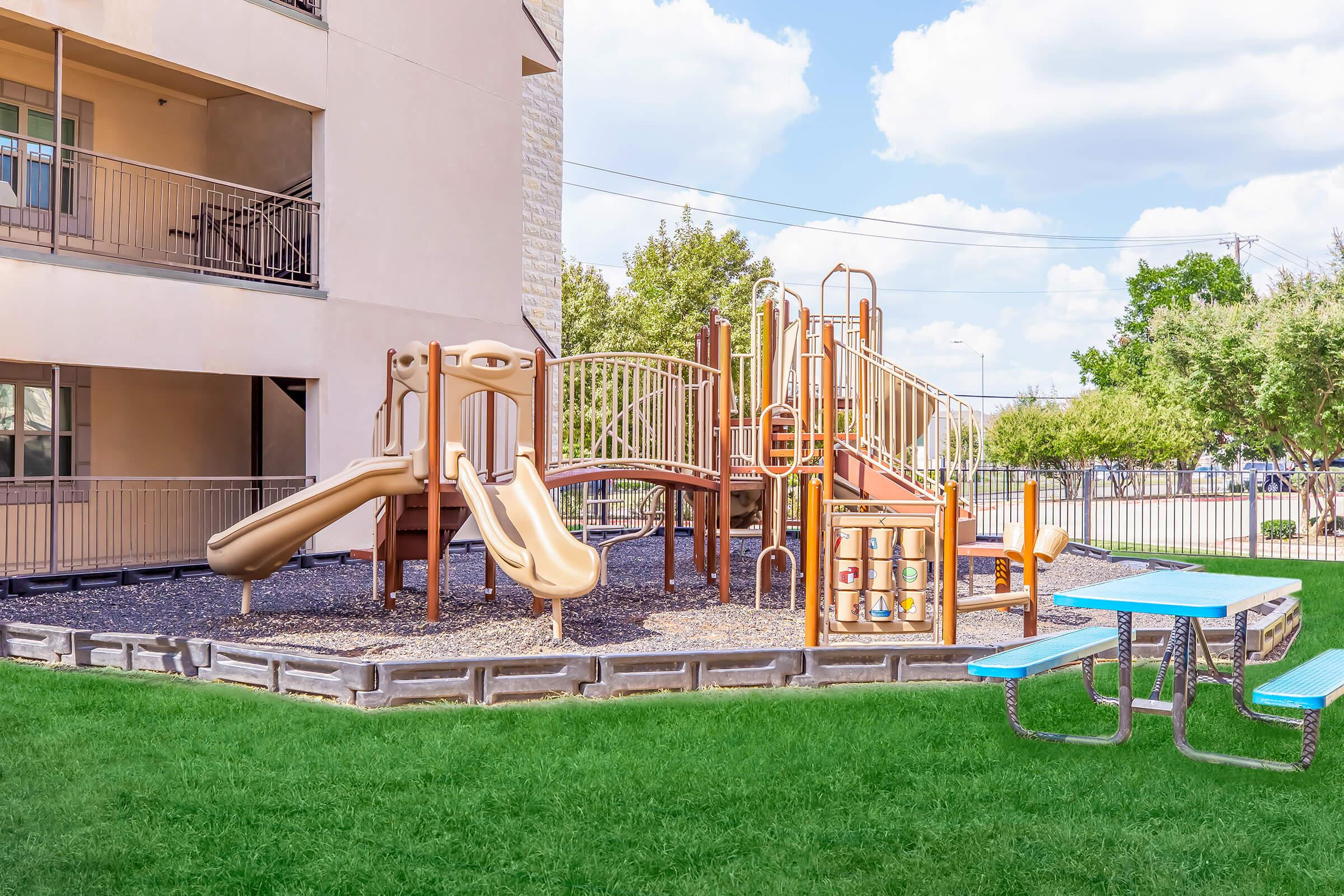 A colorful playground featuring slides and climbing structures, set on a grass area beside a building. There are picnic tables nearby, and the scene is bright with blue skies and fluffy clouds.