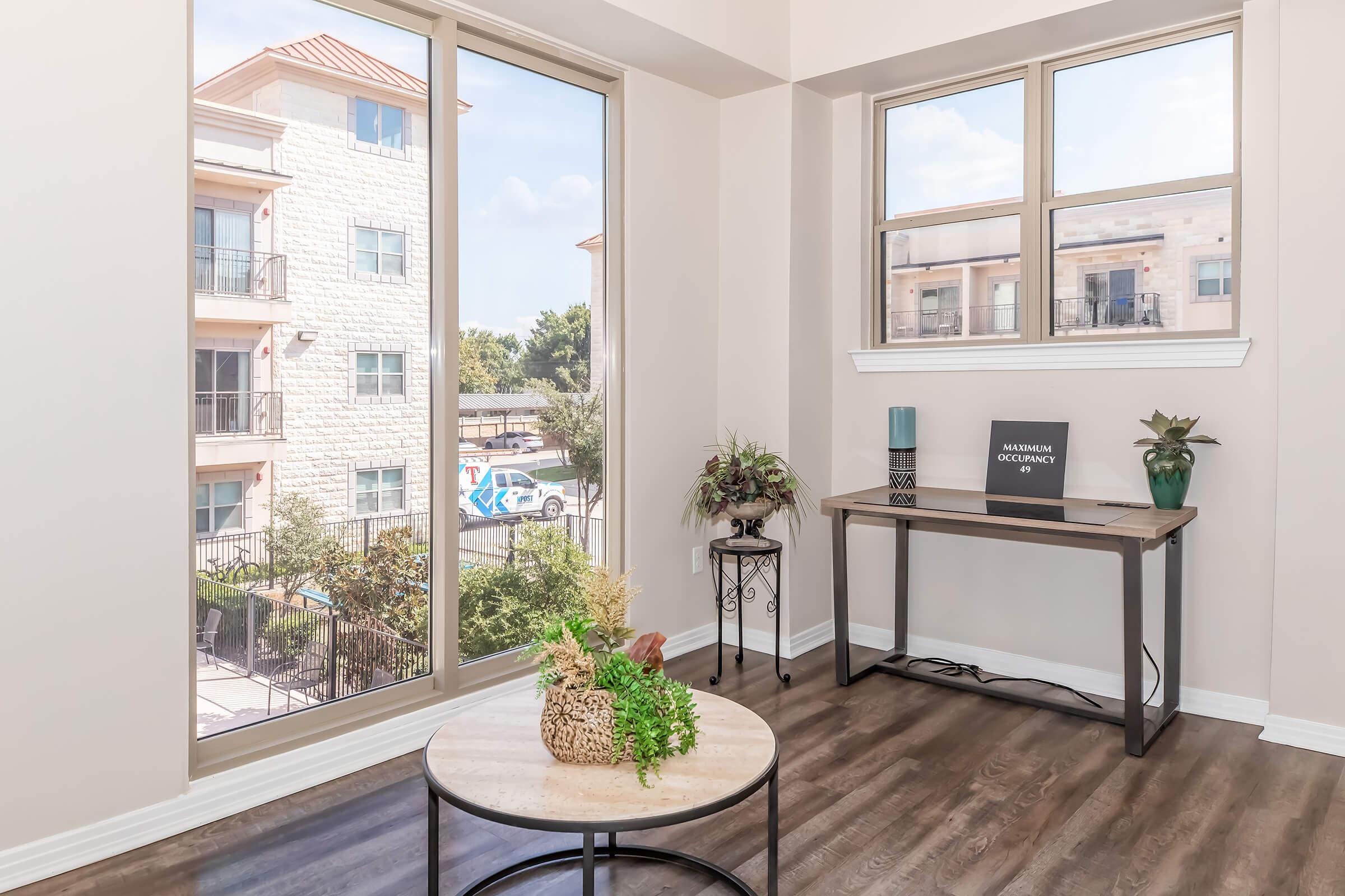 Bright interior of a modern room featuring large windows with a view of a courtyard. A wooden table with decorative plants and a sign reading "Modern Living" sits against the wall. The flooring is dark wood, and there are light-colored walls enhancing the spacious feel.