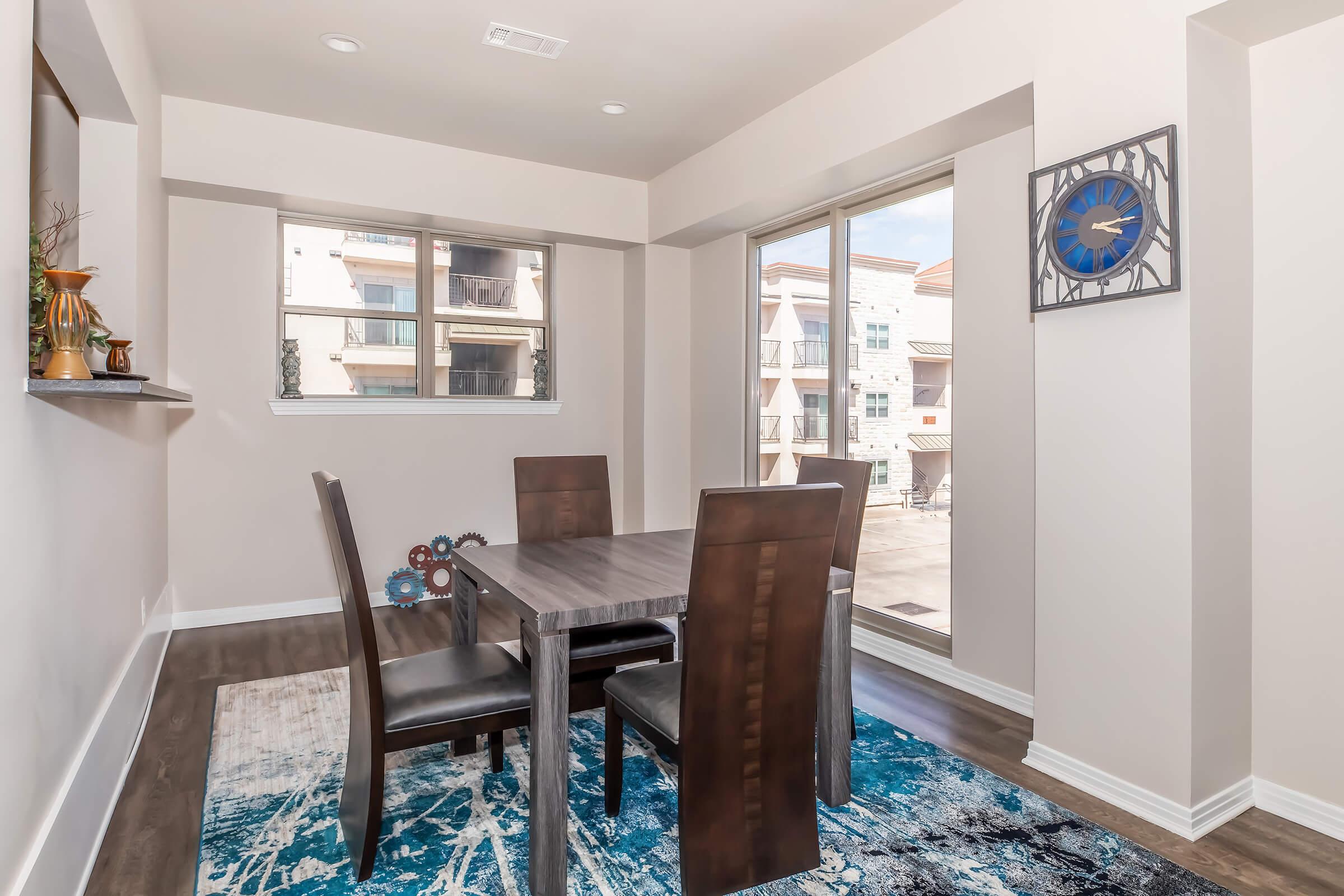 A modern dining area featuring a rectangular wooden table surrounded by four leather chairs. A decorative blue and white rug is beneath the table, and large windows allow natural light to fill the space. A clock is mounted on the wall, and a small shelf displays decorative items.