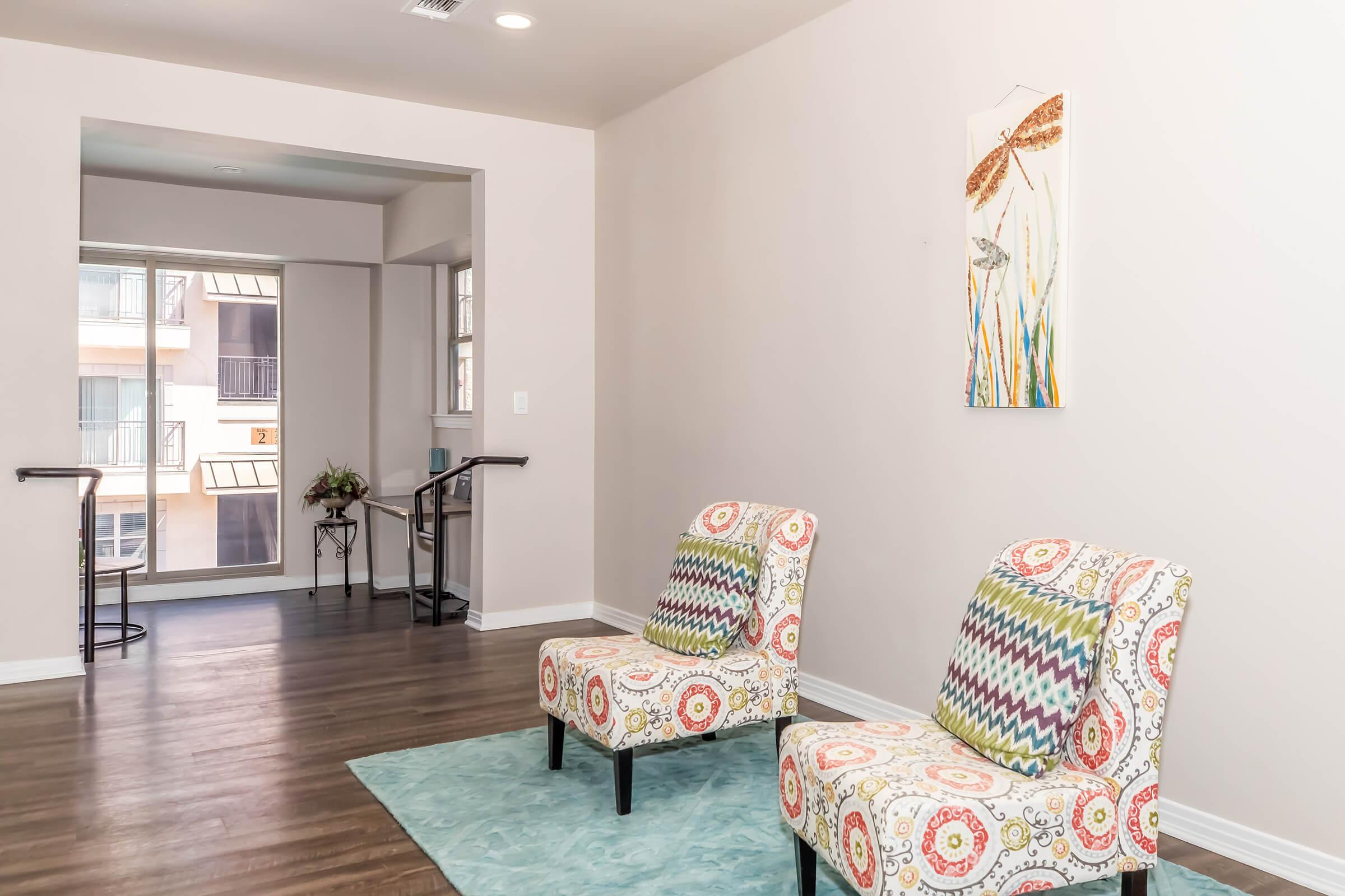 A modern interior space featuring two patterned chairs on a light blue rug. The room has light-colored walls and a welcoming atmosphere, with a small table in the corner and a window displaying natural light. A wall decoration with floral elements adds a touch of color.