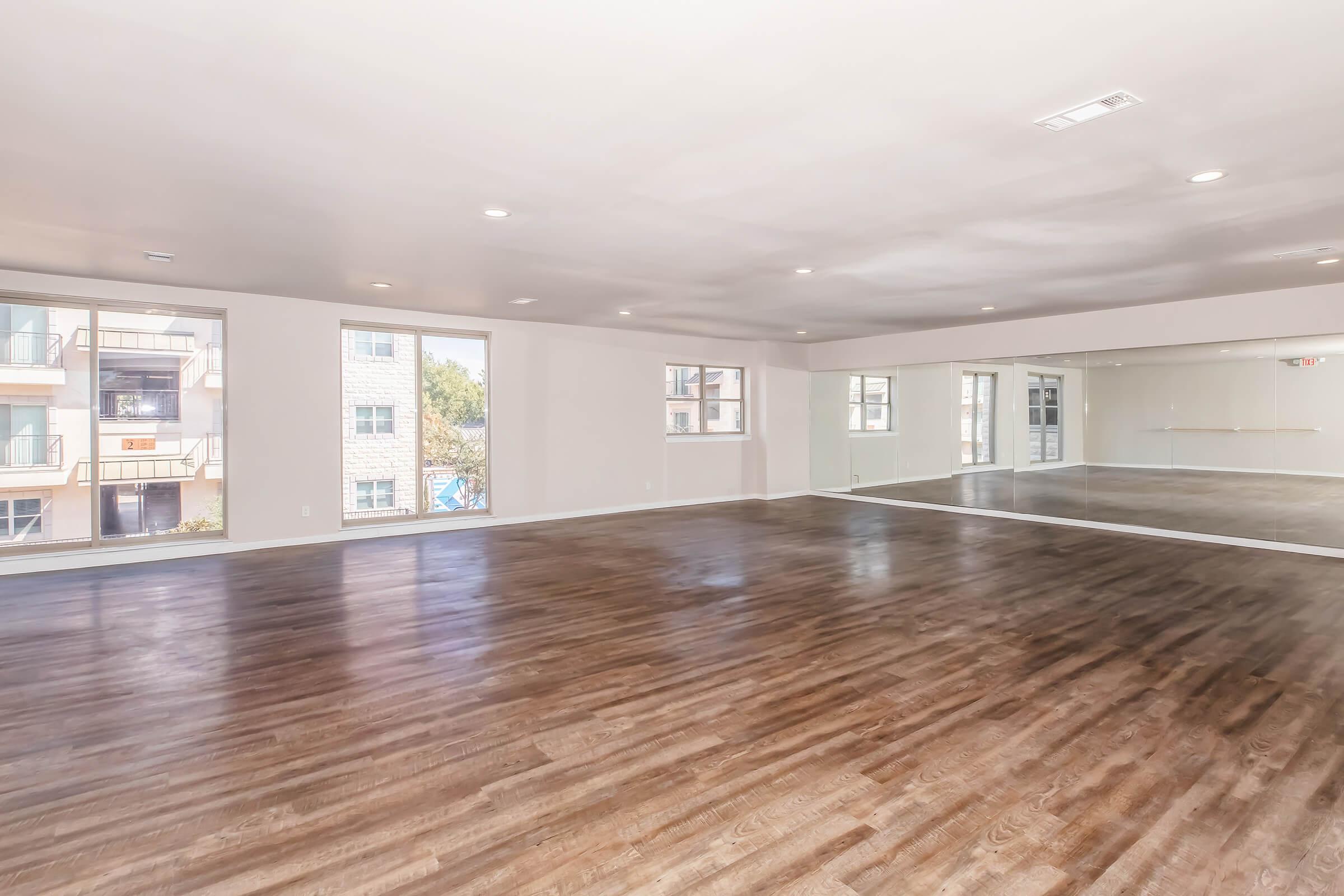 Spacious, empty room with wooden flooring, large windows allowing natural light, and a mirrored wall. The walls are painted in a light color, creating an airy atmosphere. There is a view of an outdoor balcony area from the windows. Ideal for activities like dance or fitness classes.