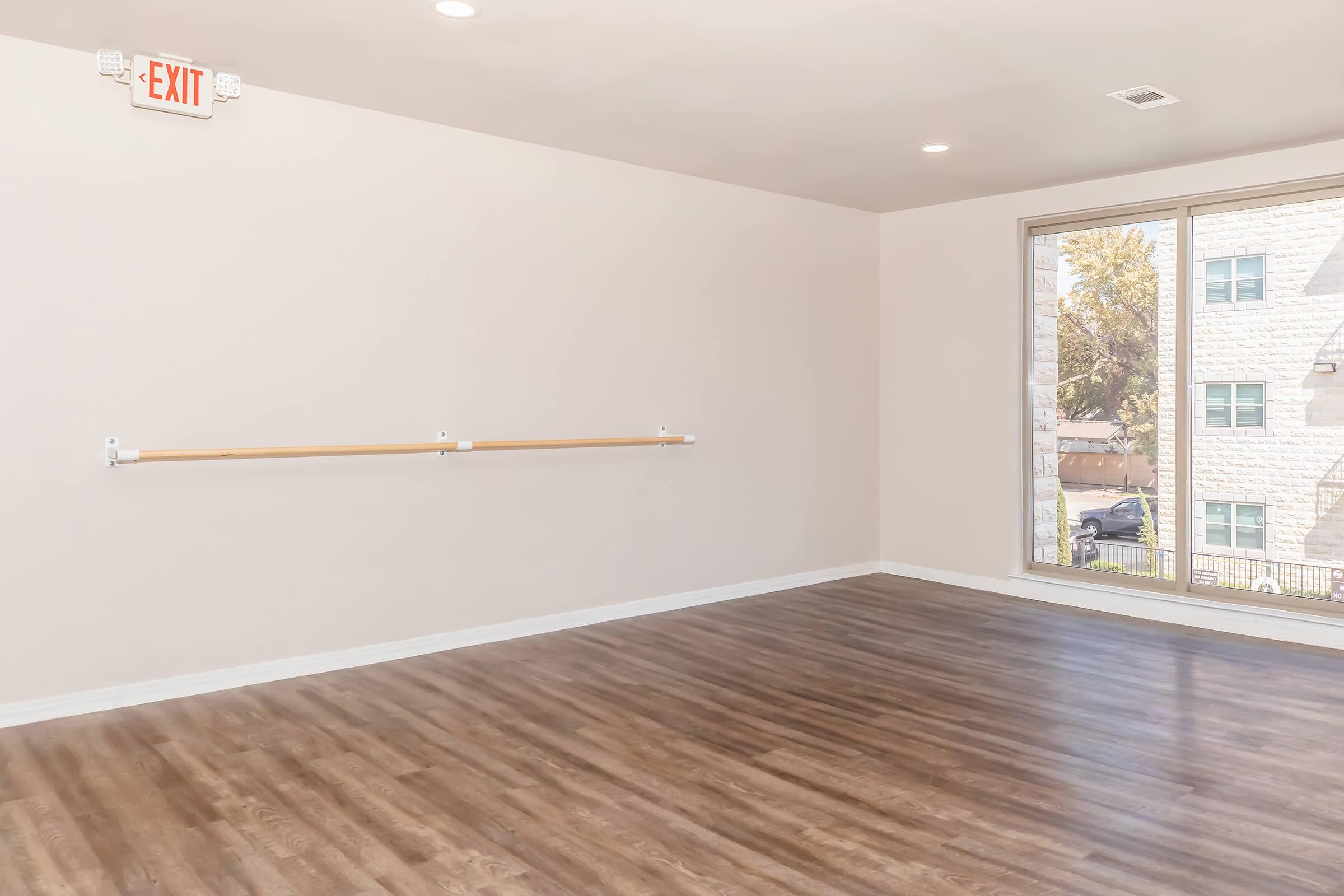 A spacious room with wooden flooring, featuring large windows that allow natural light to enter. One wall has a wooden ballet bar mounted, and an exit sign is visible on the ceiling. The walls are painted in a neutral color, and there's a view of an outdoor area through the windows.