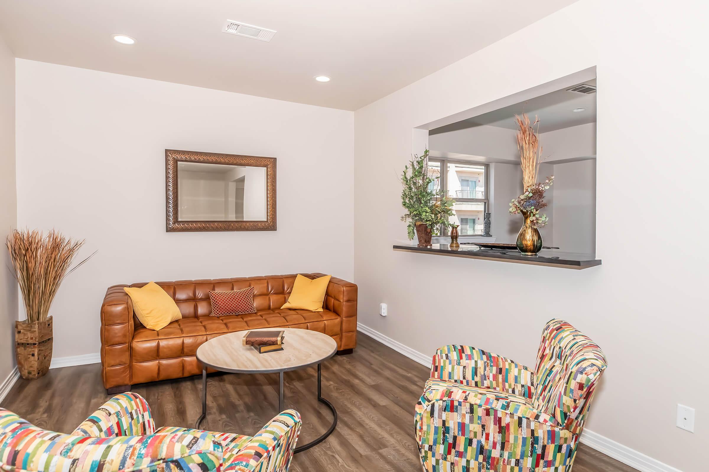 Cozy living space featuring a brown leather couch adorned with yellow throw pillows, two colorful patterned chairs, a round coffee table, and a large mirror. Decorative plants are placed in a corner and on a shelf behind the couch, with soft lighting accentuating the warm atmosphere.