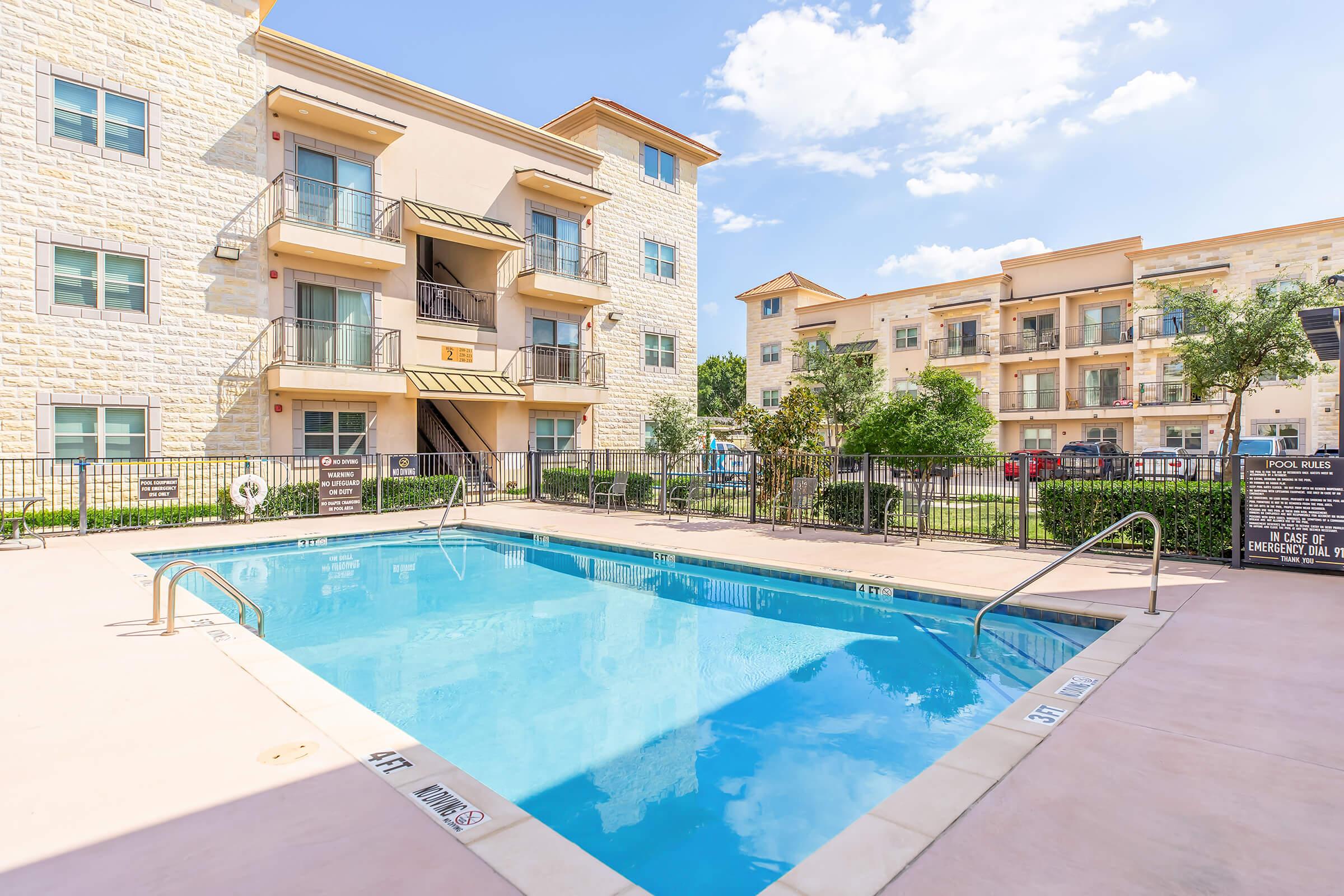 Swimming pool with clear blue water surrounded by a light-colored pavement. In the background, there are two multi-story residential buildings with light stone facades and balcony access. The sky is partly cloudy, and there are a few trees and shrubs around the pool area.