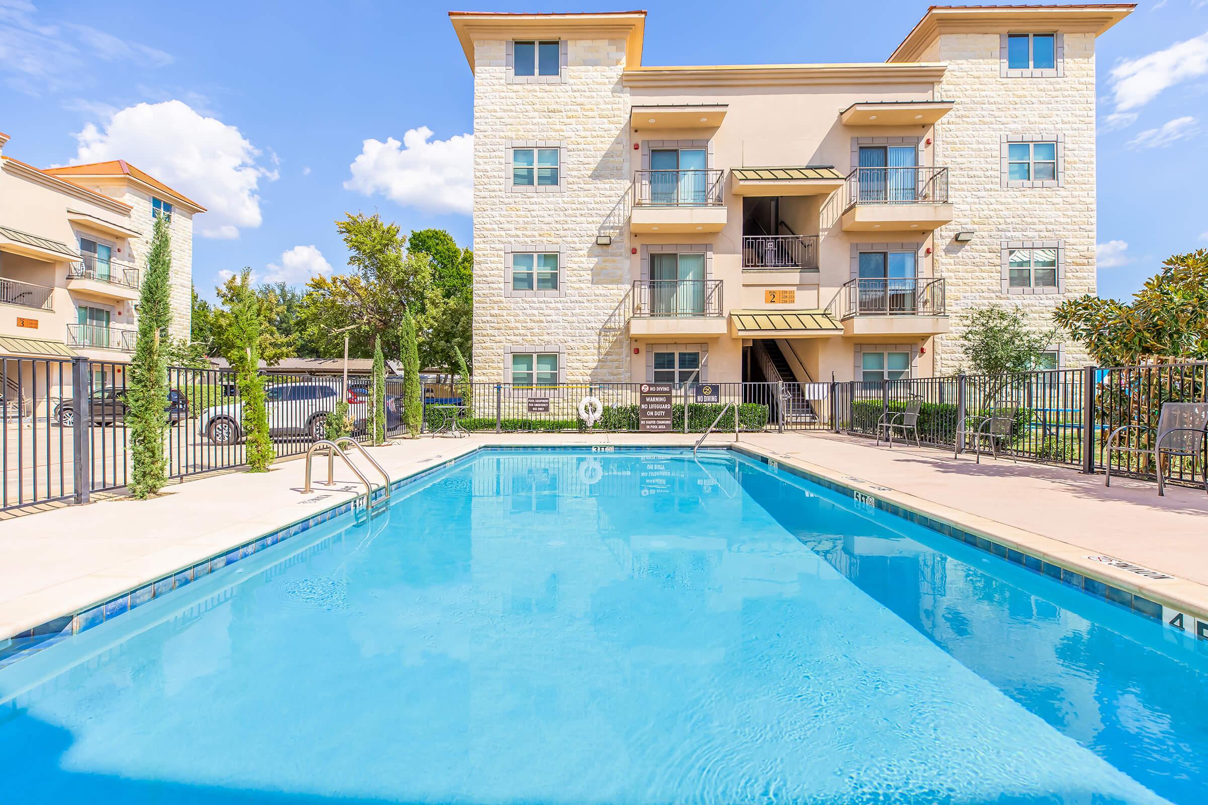 A clear blue swimming pool surrounded by a fence, with an apartment building in the background. The building features three stories with balconies, and there are trees and greenery nearby. The sky is sunny with a few clouds.