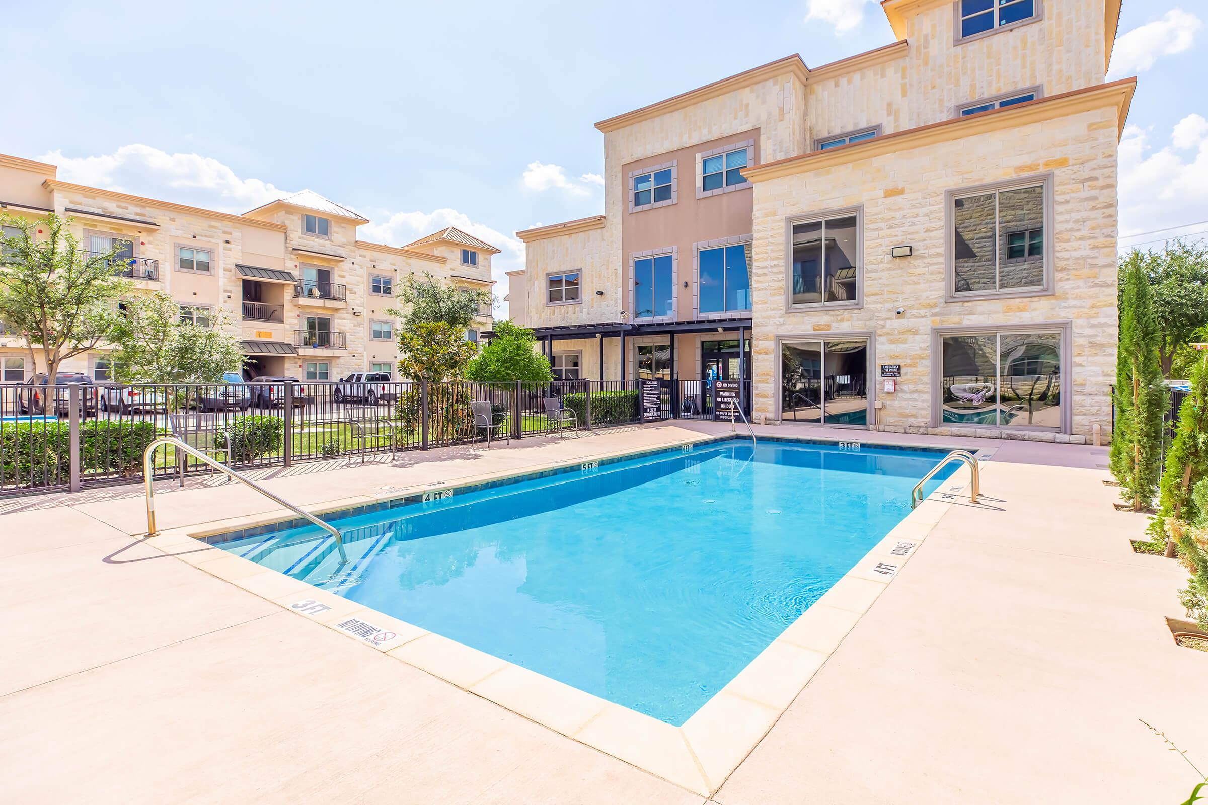 A clear blue swimming pool surrounded by a well-maintained patio area, with a few lounge chairs nearby. In the background, there are modern apartment buildings with large windows and greenery, under a sunny sky with a few clouds. The area is inviting and well-designed for relaxation.
