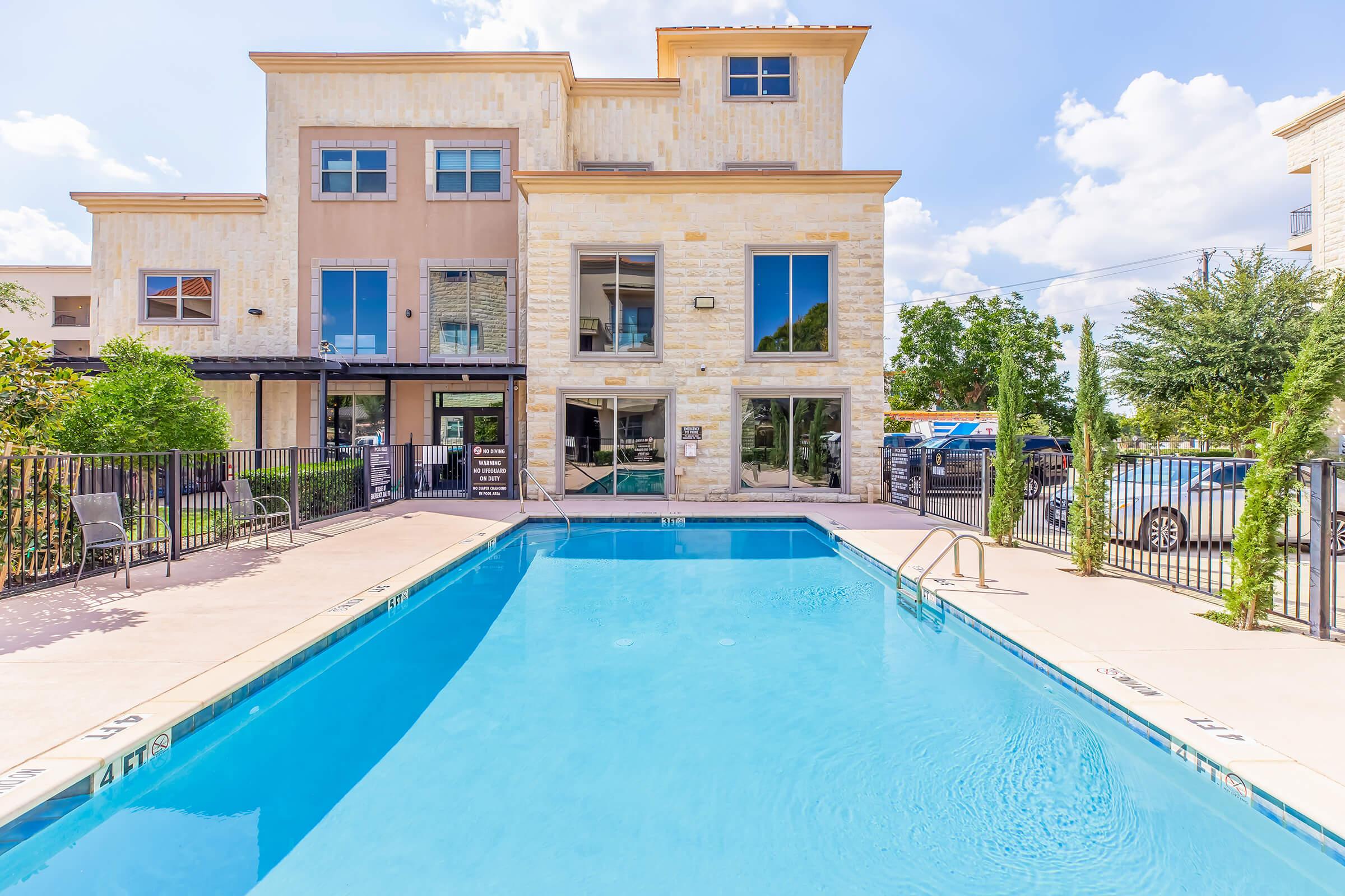A clear swimming pool surrounded by a patio area, with lounge chairs on one side. In the background, there is a modern building with large windows and a mix of stone and siding exterior. Trees and greenery frame the scene under a bright blue sky with fluffy clouds.