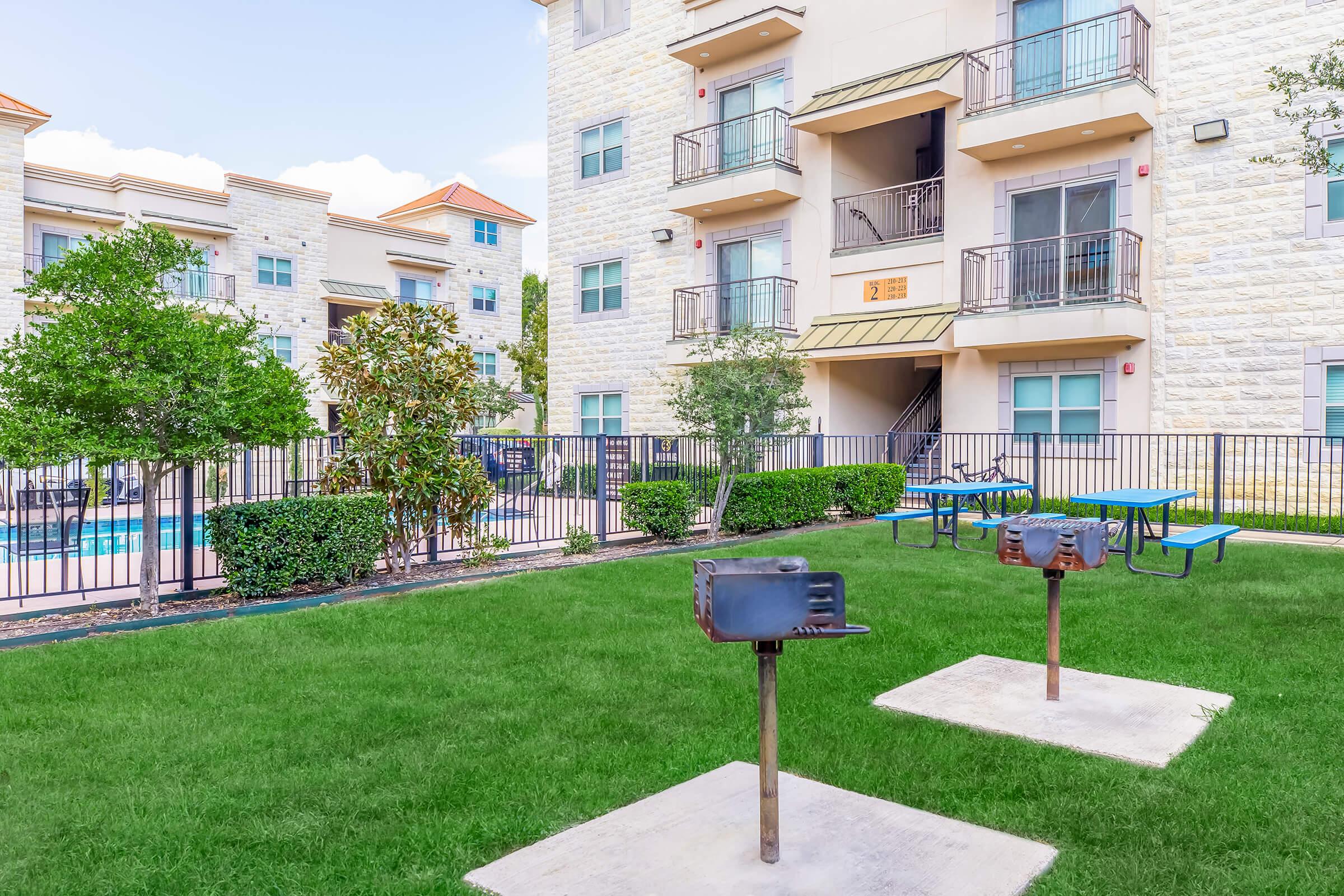 A well-maintained apartment complex featuring a green lawn with two barbecue grills on concrete pads. In the background, there are several buildings with balconies, surrounded by trees and shrubs. A swimming pool is visible behind a fence, enhancing the outdoor recreational area.