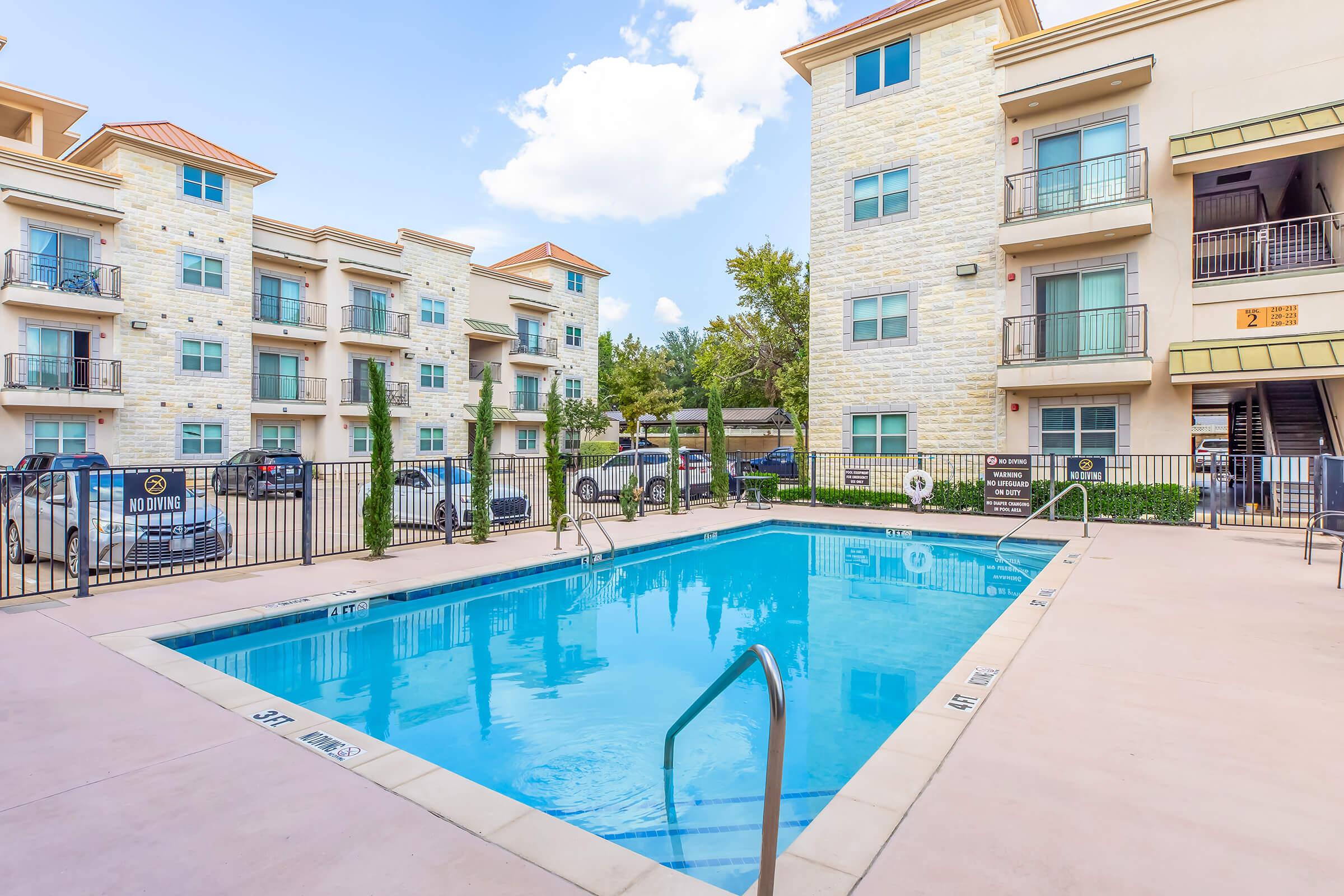 A bright outdoor pool surrounded by an apartment complex. The pool features clear blue water and steps for easy entry, with lounge chairs and a fenced area. The buildings are made of light-colored stone, with balconies overlooking the pool area, under a partly cloudy sky.