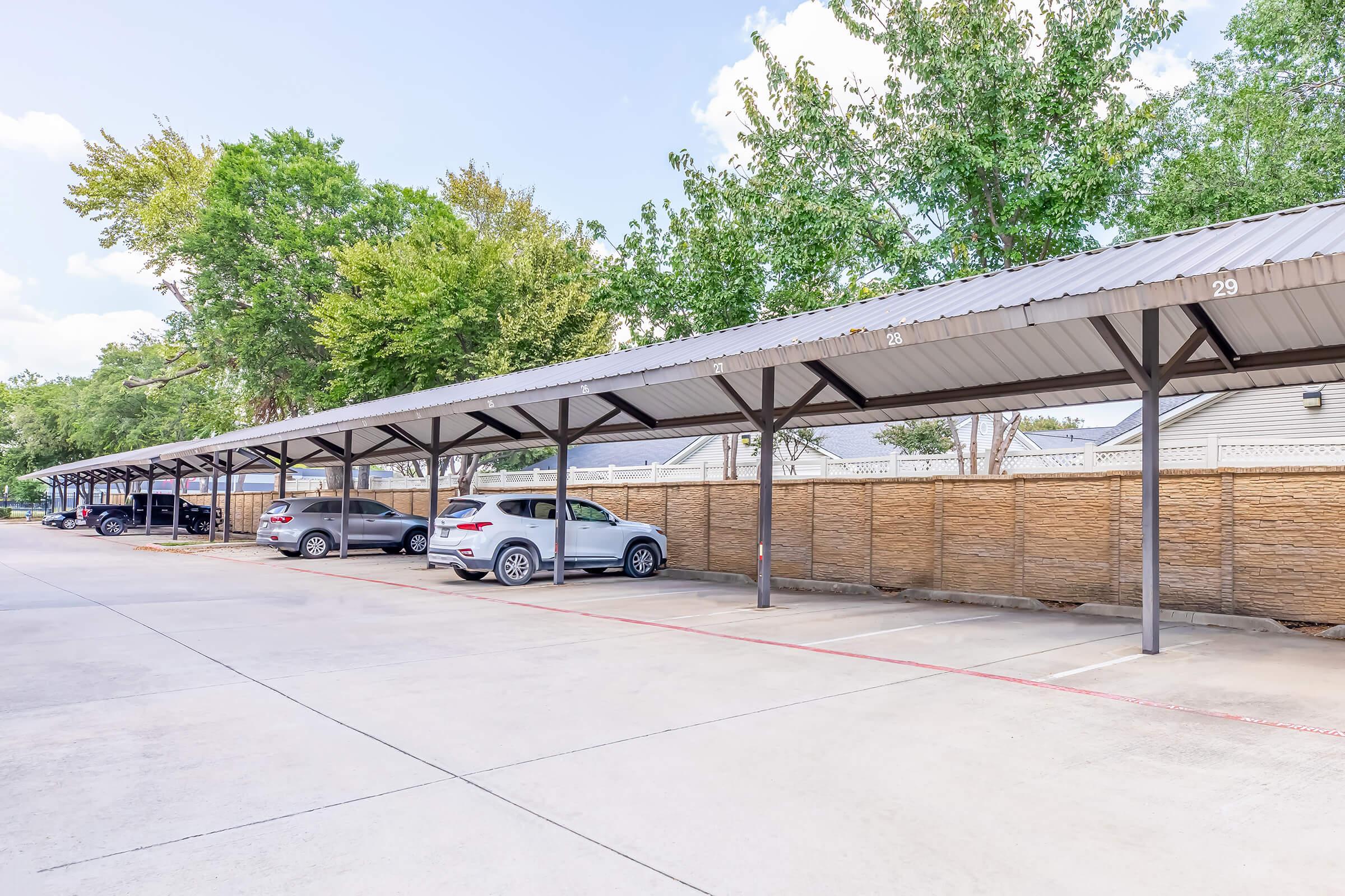 A covered parking area featuring multiple parking spaces, some occupied by cars. The structure has a metal roof supported by posts, with trees and a fence visible in the background. The scene is set on a clear day with blue skies and scattered clouds.