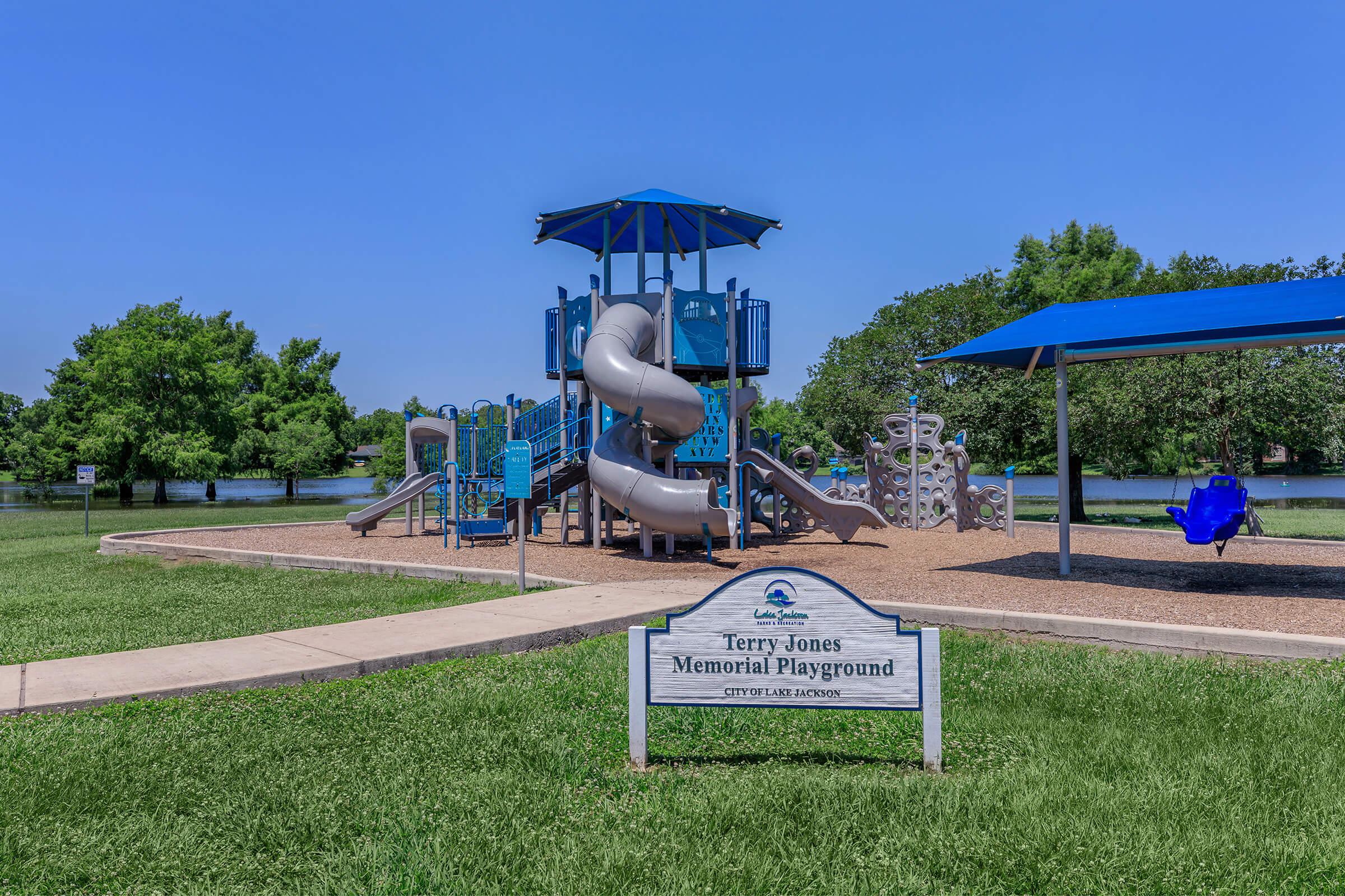 A playground featuring a large gray slide, climbing structures, and blue canopies, situated in a grassy area near a body of water. A sign reading "Terry Jones Memorial Playground" is visible in front of the playground.