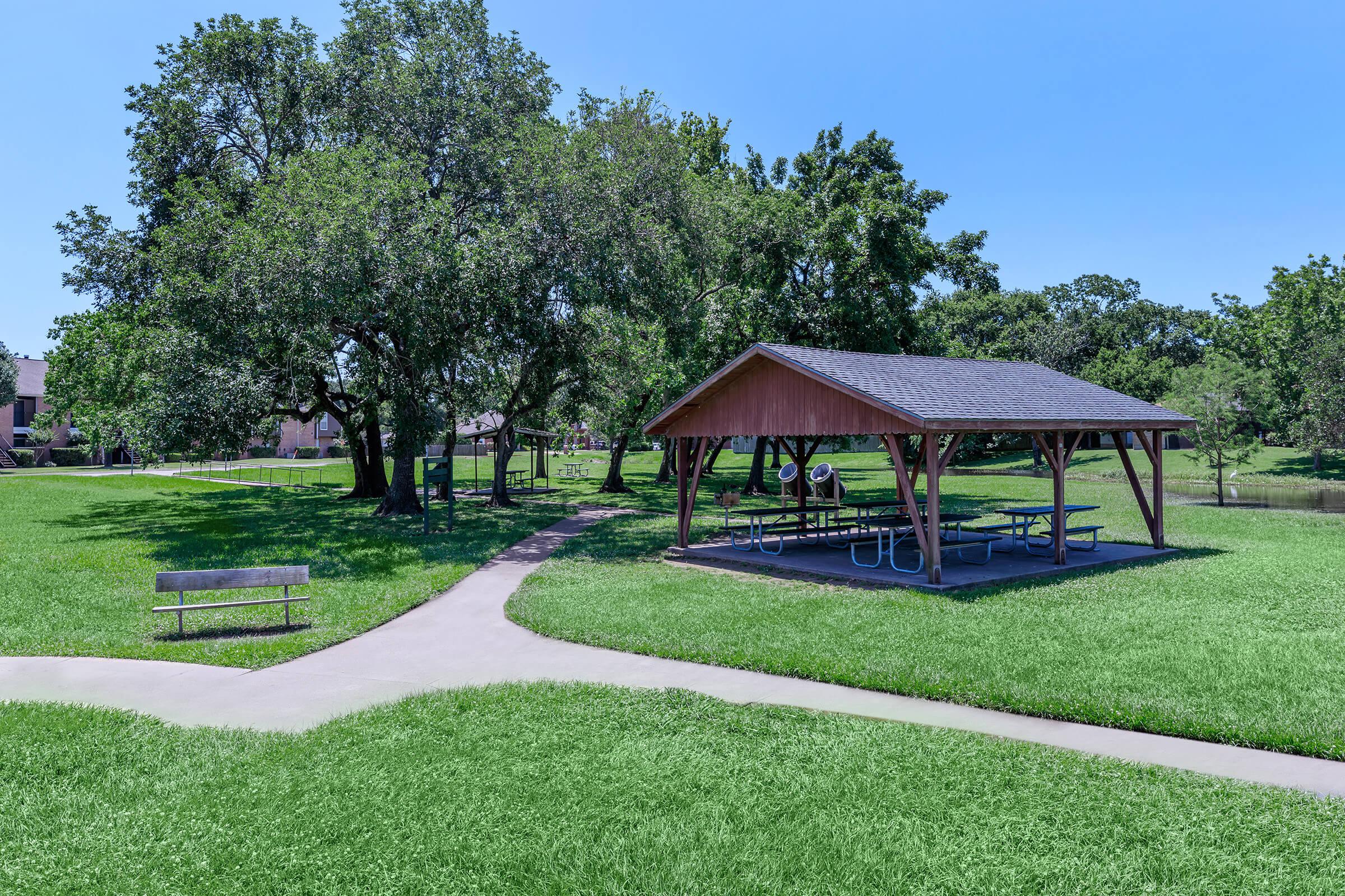 A sunny park scene featuring a wooden pavilion with picnic tables, surrounded by green grass and large trees. A pathway winds through the park, leading to various areas, while a bench is placed nearby.