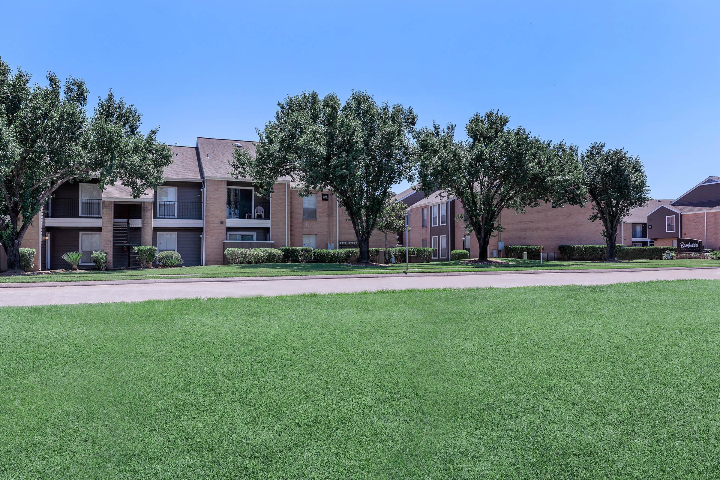 A clear, sunny day view of a residential area featuring two-story brick apartment buildings surrounded by well-maintained grassy lawns and trees.