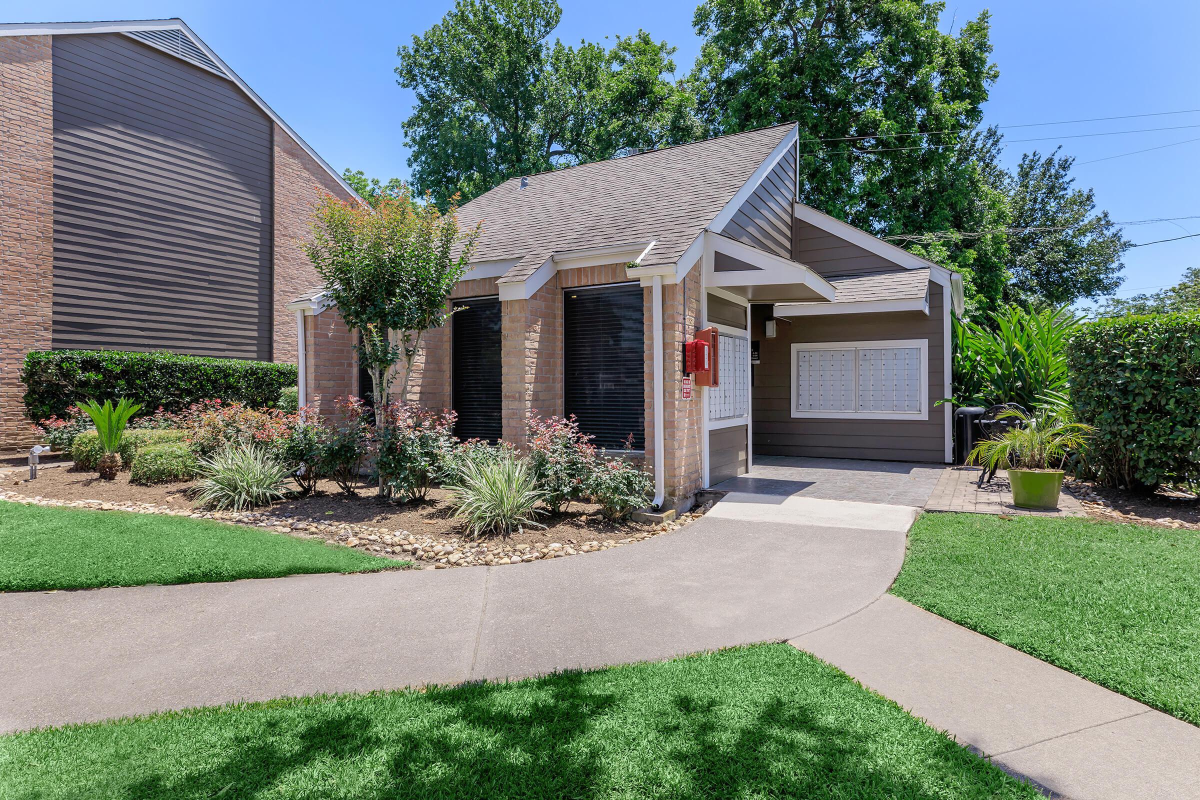 A well-maintained single-story house surrounded by lush greenery and landscaped gardens. The pathway leads to the entrance, with colorful plants and a mailbox visible. The exterior features a blend of brick and siding, under a clear blue sky.