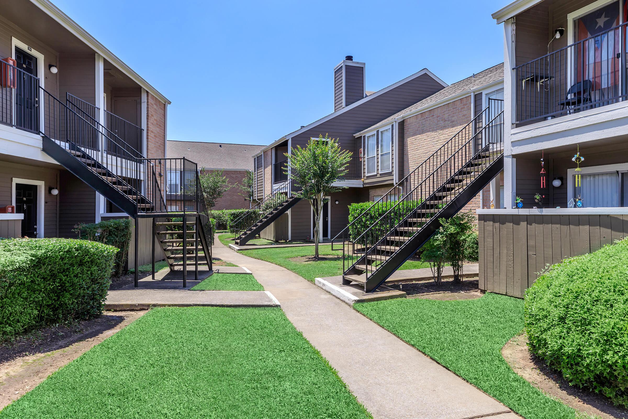 View of an apartment community courtyard featuring two-story buildings with balconies, surrounded by manicured green lawns and small trees. A concrete path runs through the middle, with staircases leading to the upper levels of the buildings. The sky is clear and blue.