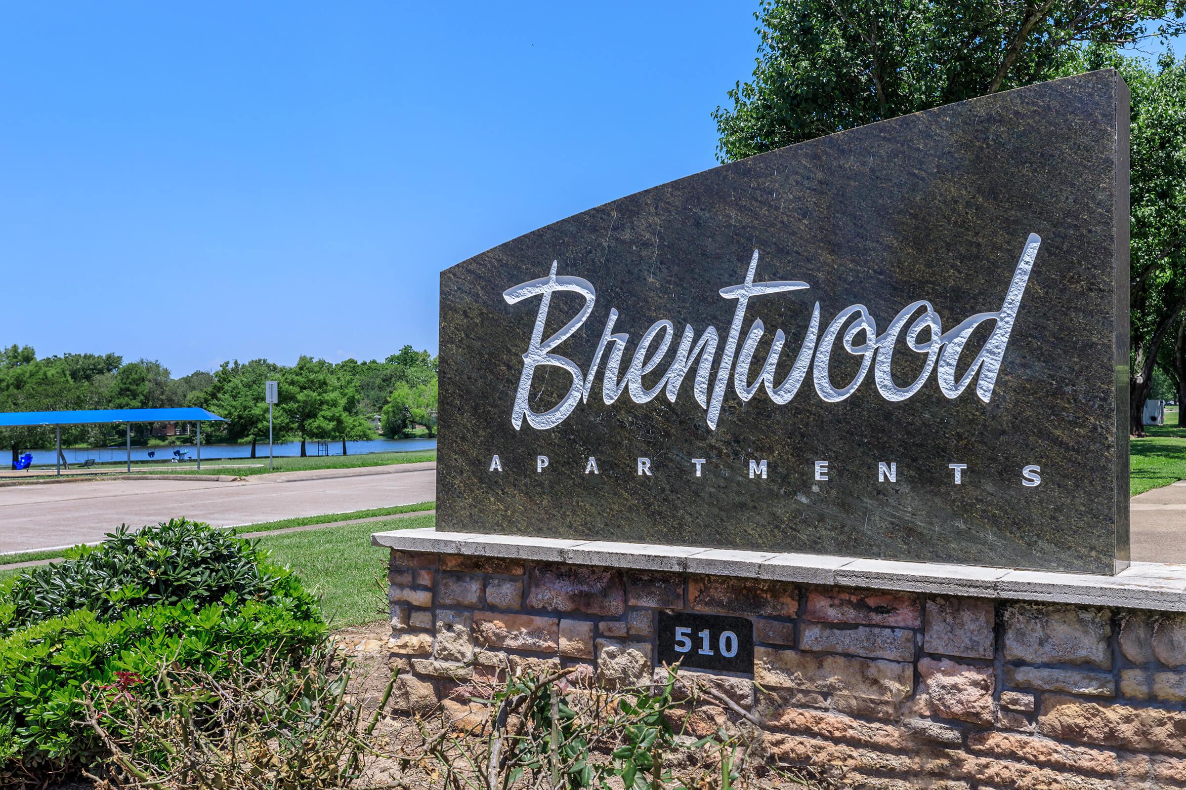 Sign for Brentwood Apartments, featuring stylized white lettering on a stone background, situated near a pond with greenery in the background. The sign displays the address number 510.