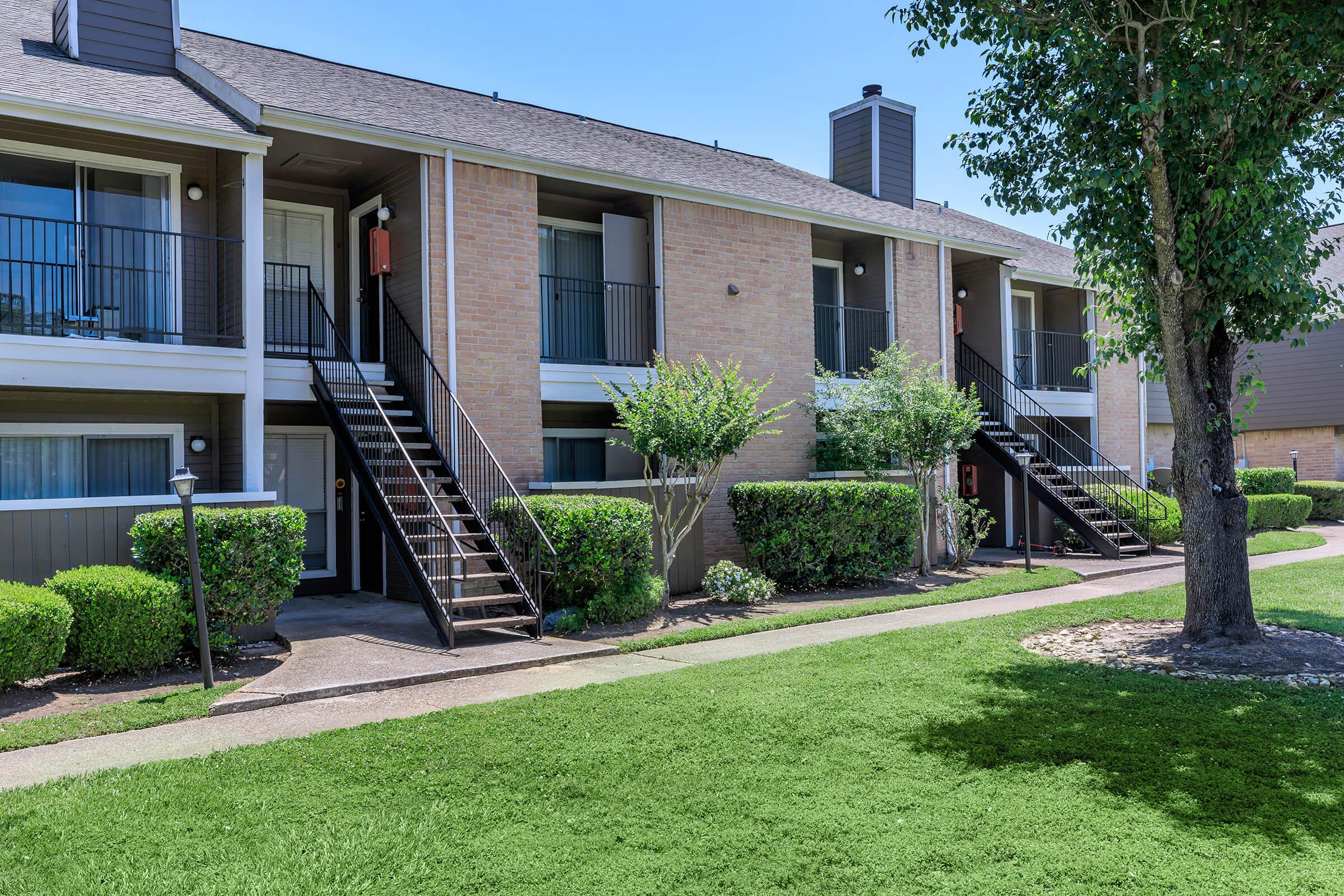 View of a two-story apartment building with a row of units, black railings, and a well-maintained lawn, featuring neatly trimmed shrubs and a tree in the foreground.