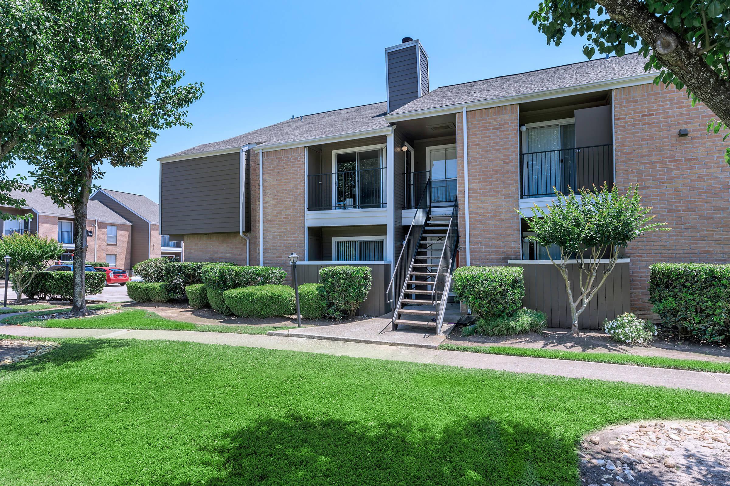 Multi-unit apartment building with brick exterior, stairs leading to second-floor units, surrounded by well-maintained greenery and lawn on a sunny day.