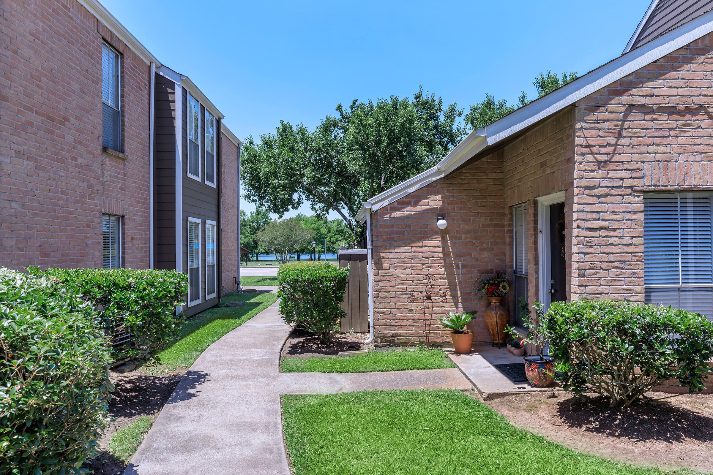 A pathway between two apartment buildings surrounded by grassy areas and bushes under a clear blue sky.