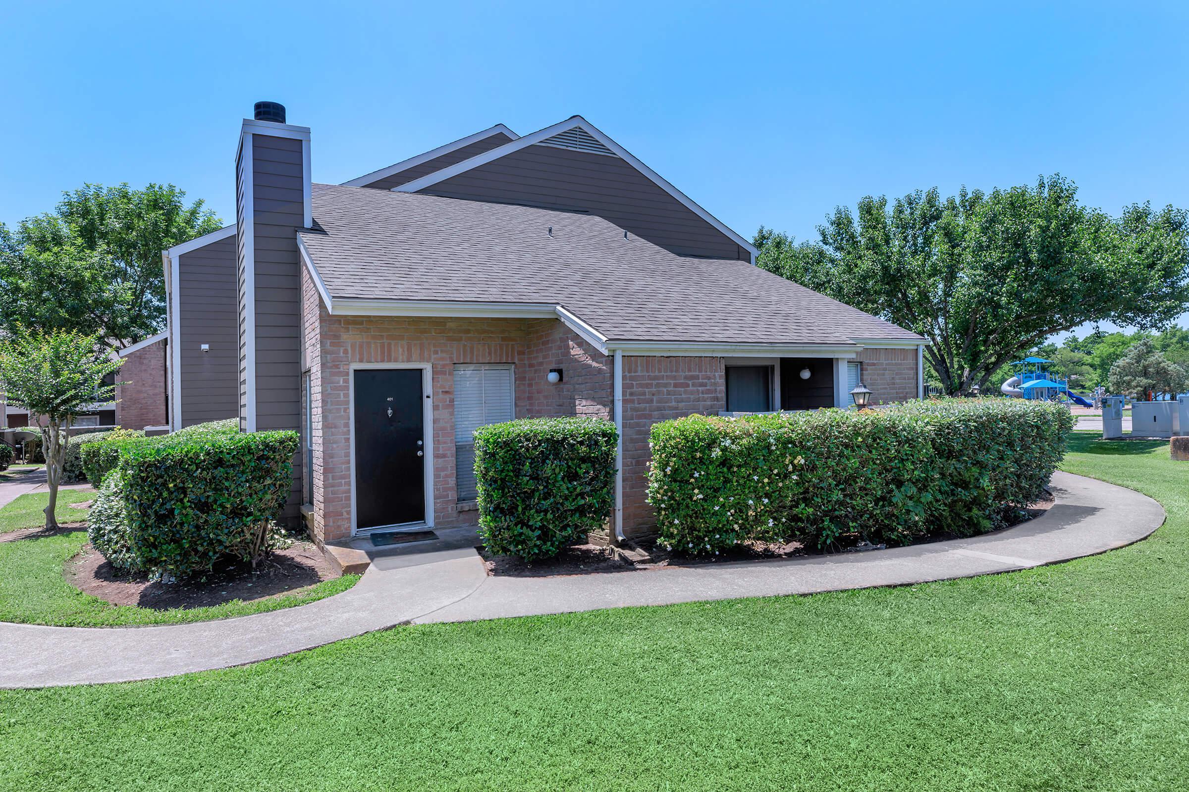 A brick apartment building with a sloped roof, surrounded by neatly trimmed hedges and green grass, under a clear blue sky.