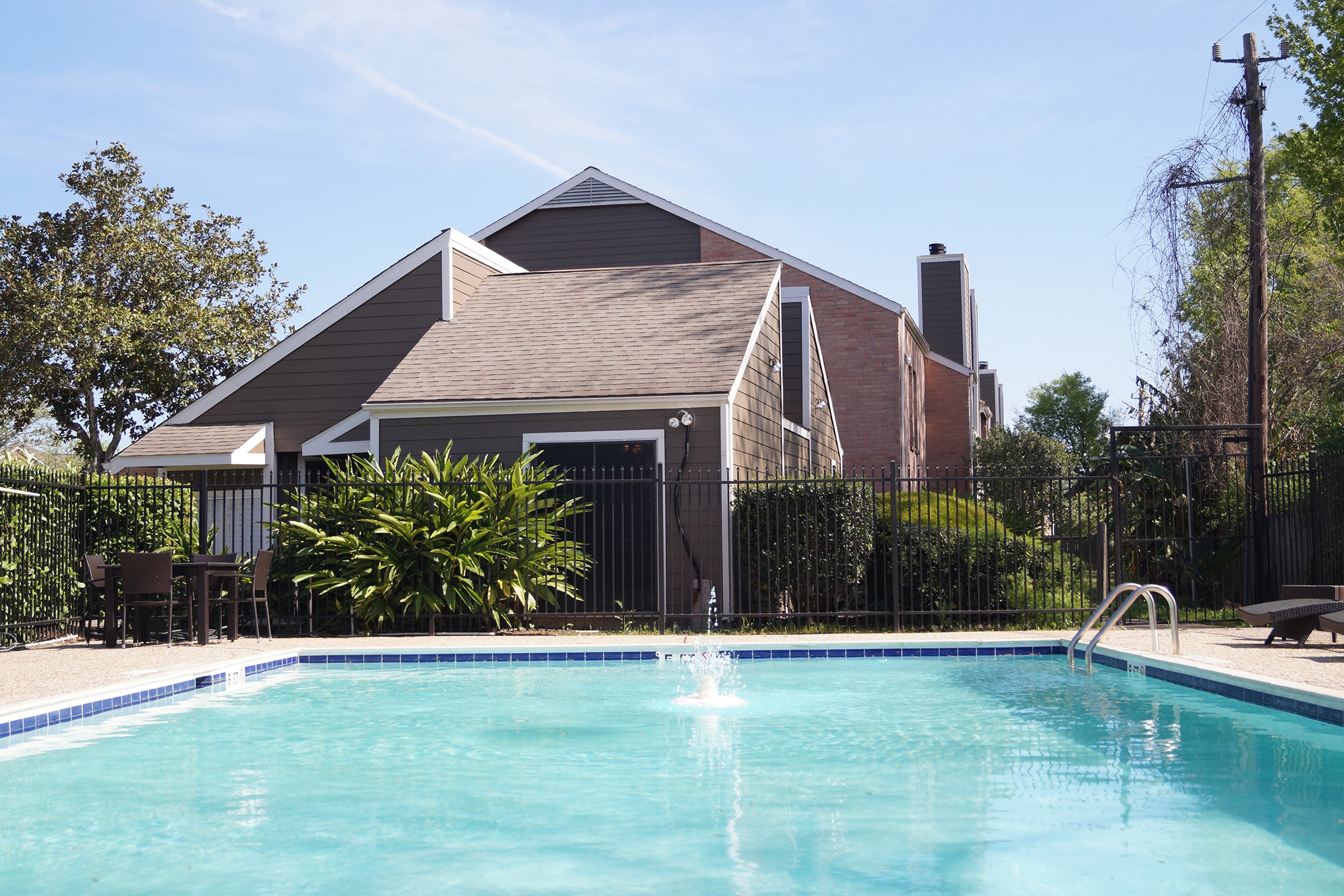 A clear swimming pool in the foreground with a fountain, surrounded by a patio area and lush greenery. In the background, there is a two-story residential building with a sloped roof and a fenced yard under a blue sky.