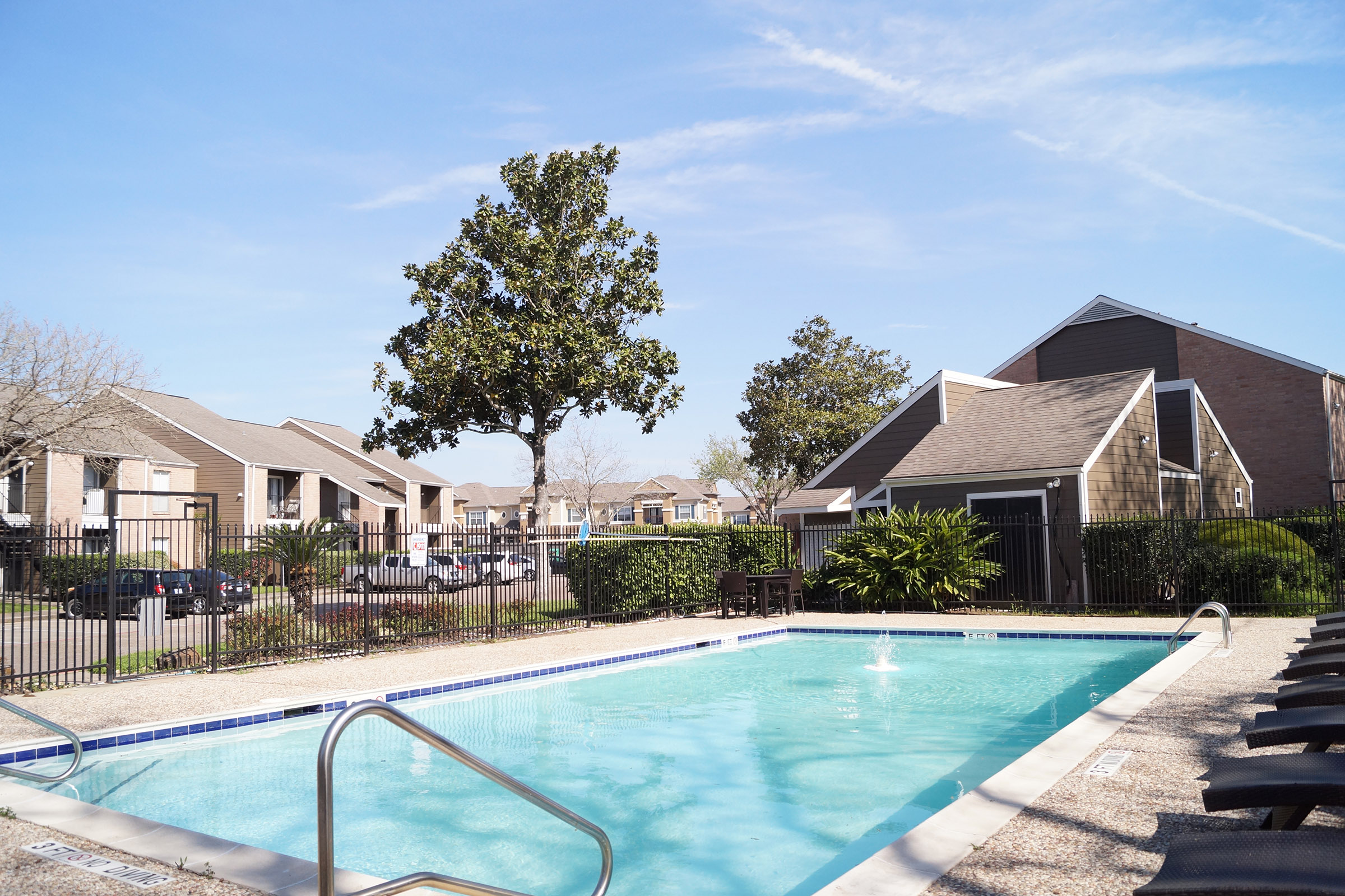 A clear blue swimming pool surrounded by lounge chairs, with nearby trees and apartment buildings in the background under a sunny sky.