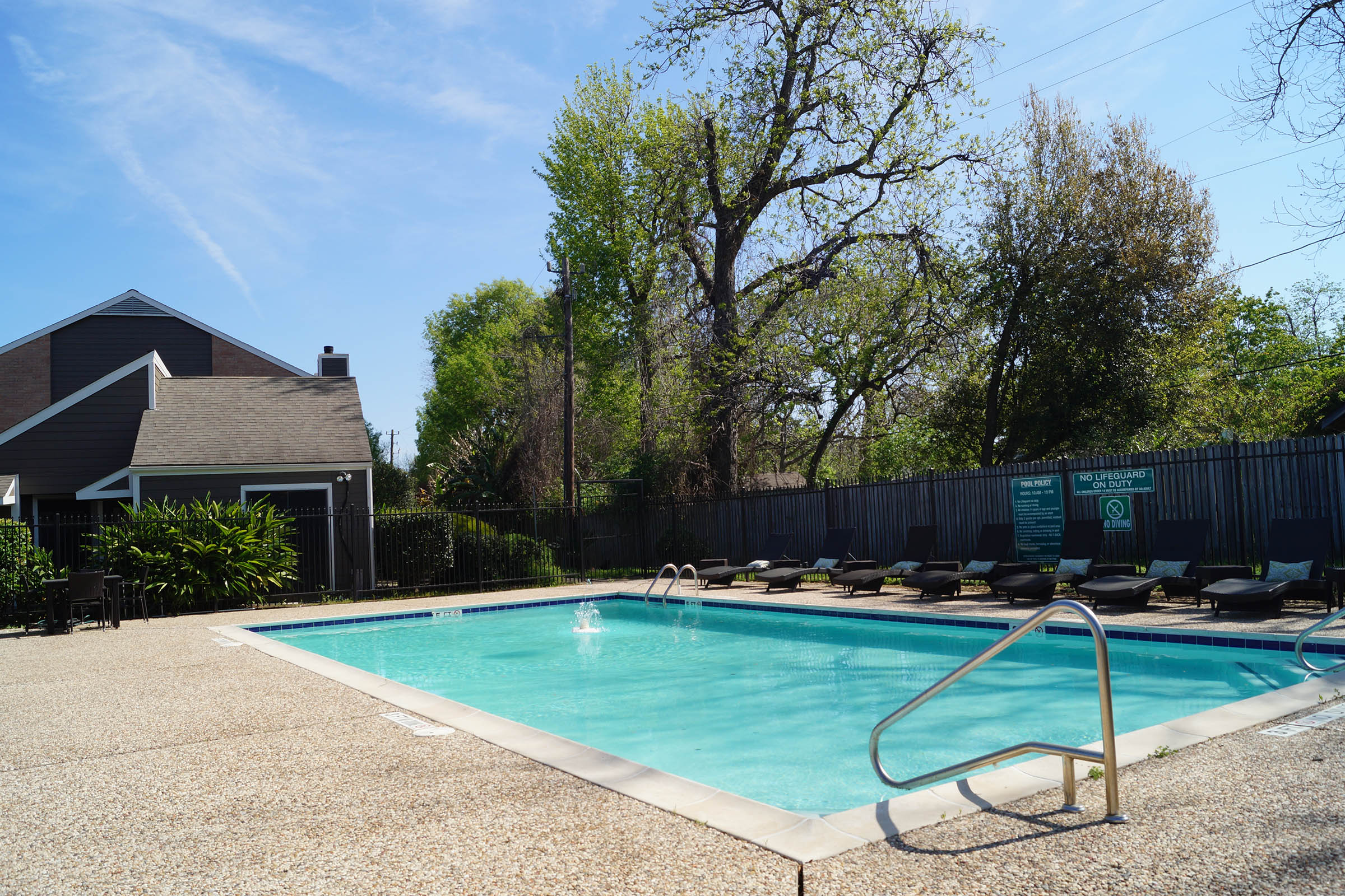 A clear blue swimming pool surrounded by lounge chairs, with trees and a residential building in the background under a sunny sky.