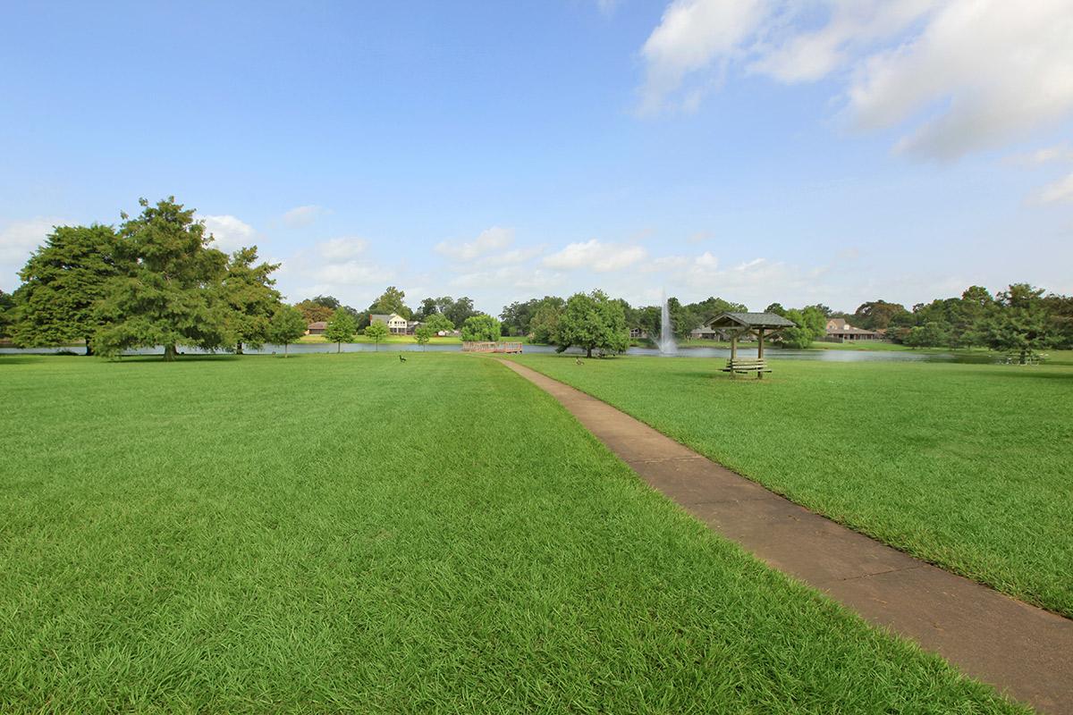 A scenic park view featuring a wide expanse of lush green grass, a concrete pathway winding through the landscape, and a gazebo in the distance. There is a small water fountain in a pond on the left side, surrounded by trees and a clear blue sky with some clouds.