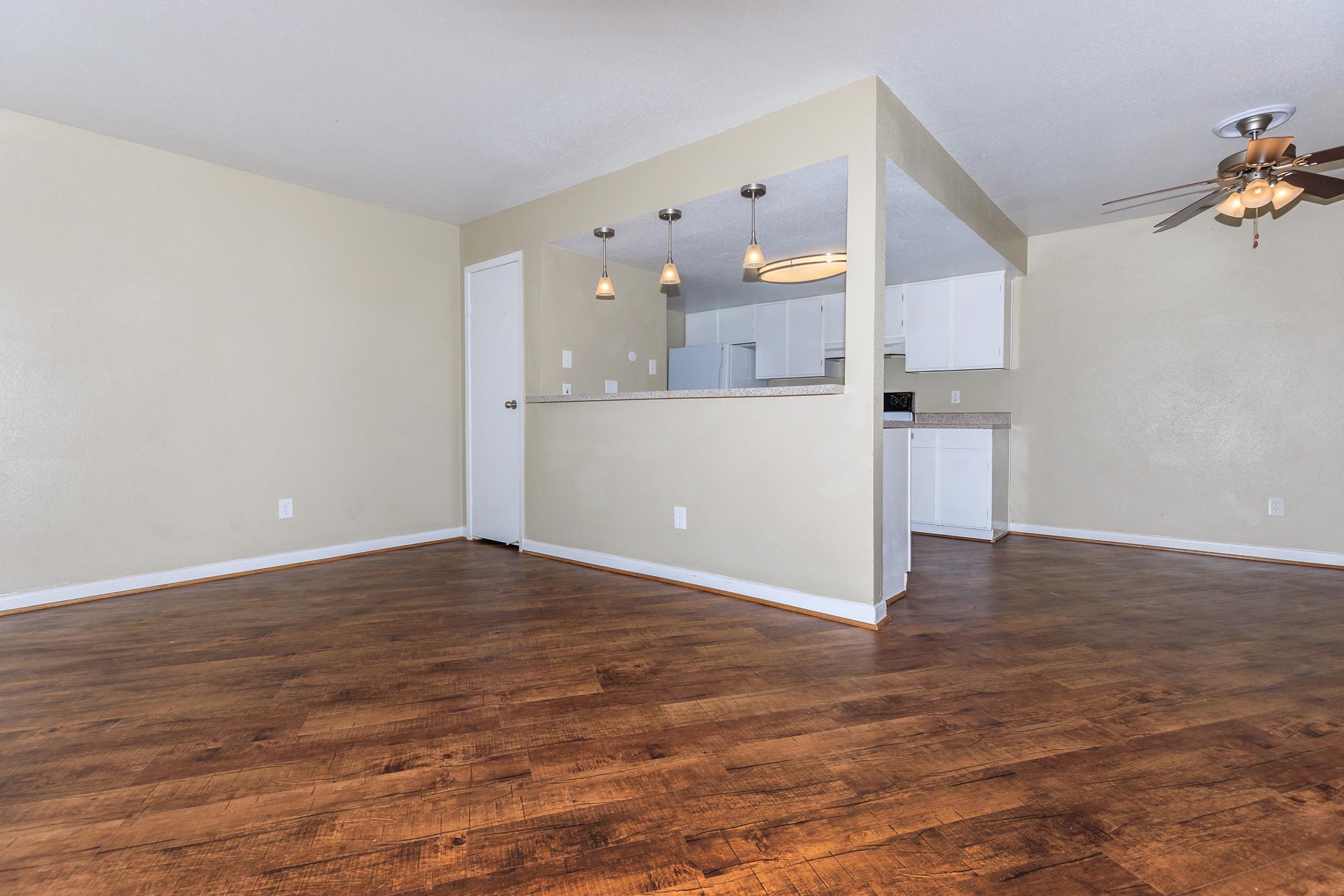 A spacious, well-lit room with wood flooring, featuring a ceiling fan. There is a doorway leading to another area and a mirrored wall that separates it from a kitchenette with white cabinets and modern light fixtures. The walls are painted light beige, creating a warm atmosphere.