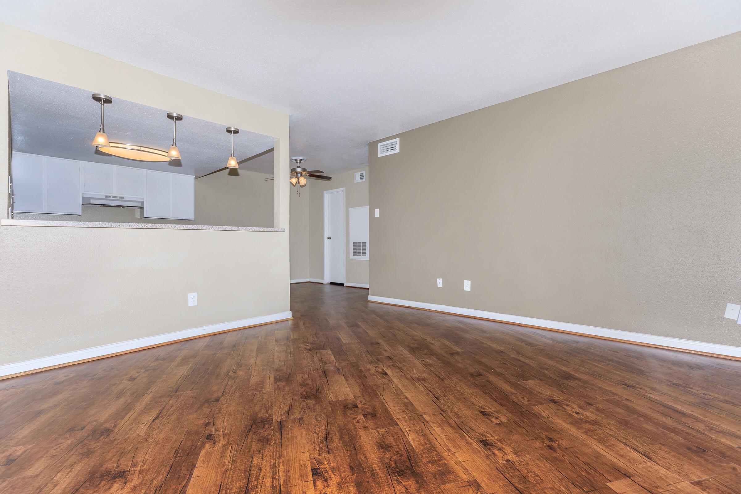 A spacious living area with hardwood floors, light-colored walls, and a ceiling fan. To the left, there is a kitchen space visible through an open area with a bar. The room is well-lit and features a doorway leading to another room.